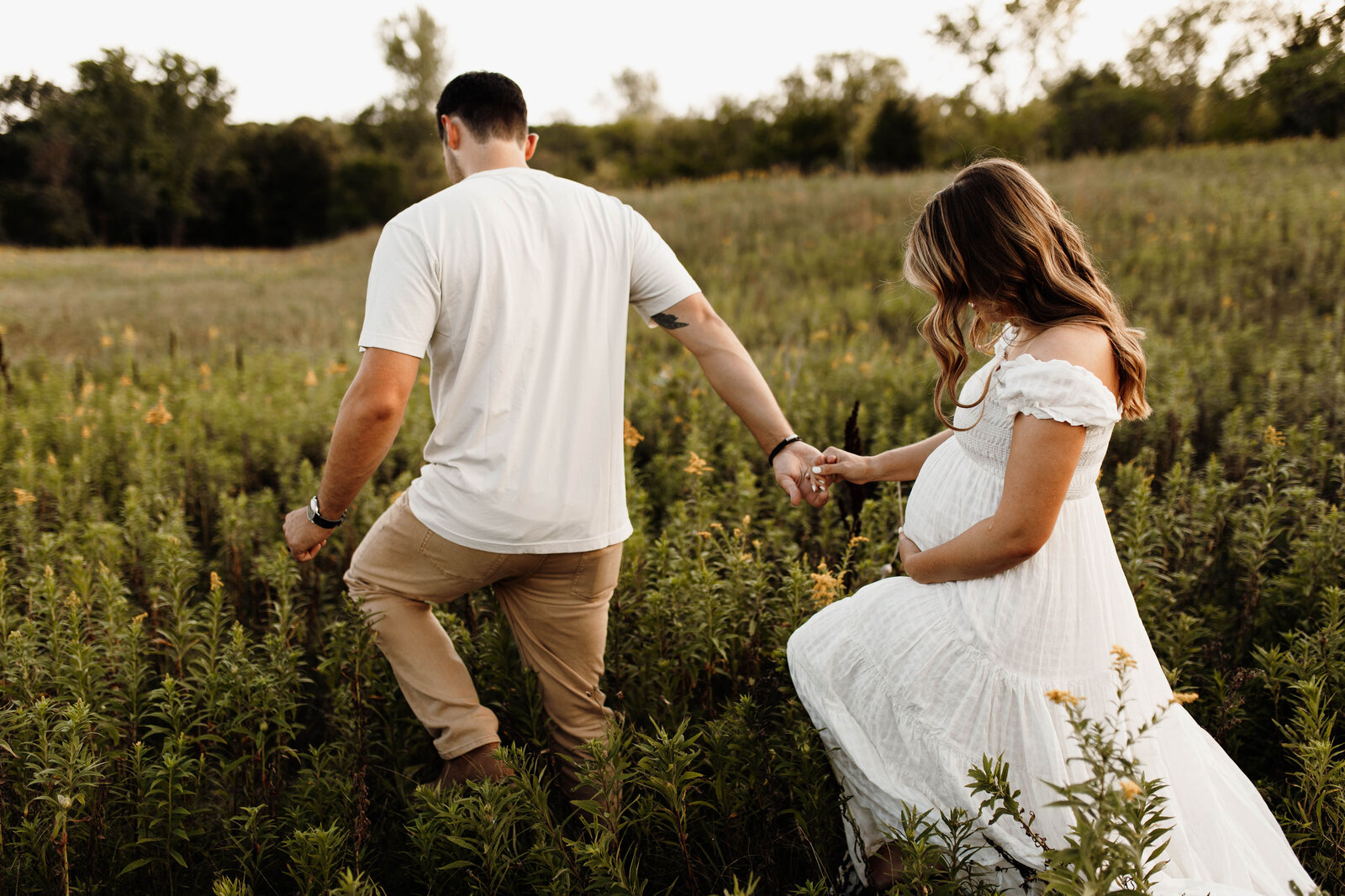 Couple holding hands and walking through a field while the woman holds her pregnant belly