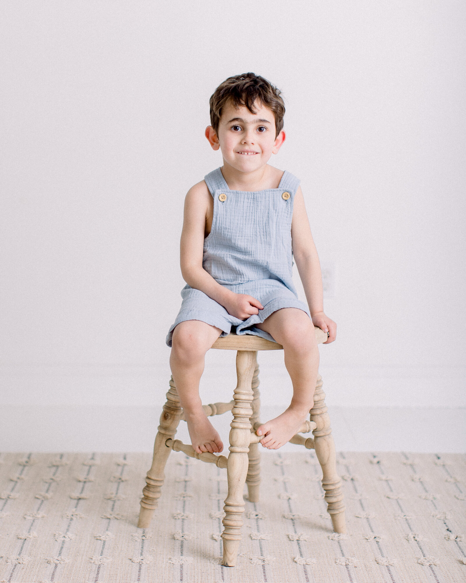 4 year old boy in blue linen overalls, sitting on a wooden stool in a neutral studio by NH newborn photographer Fieldstone Studio.