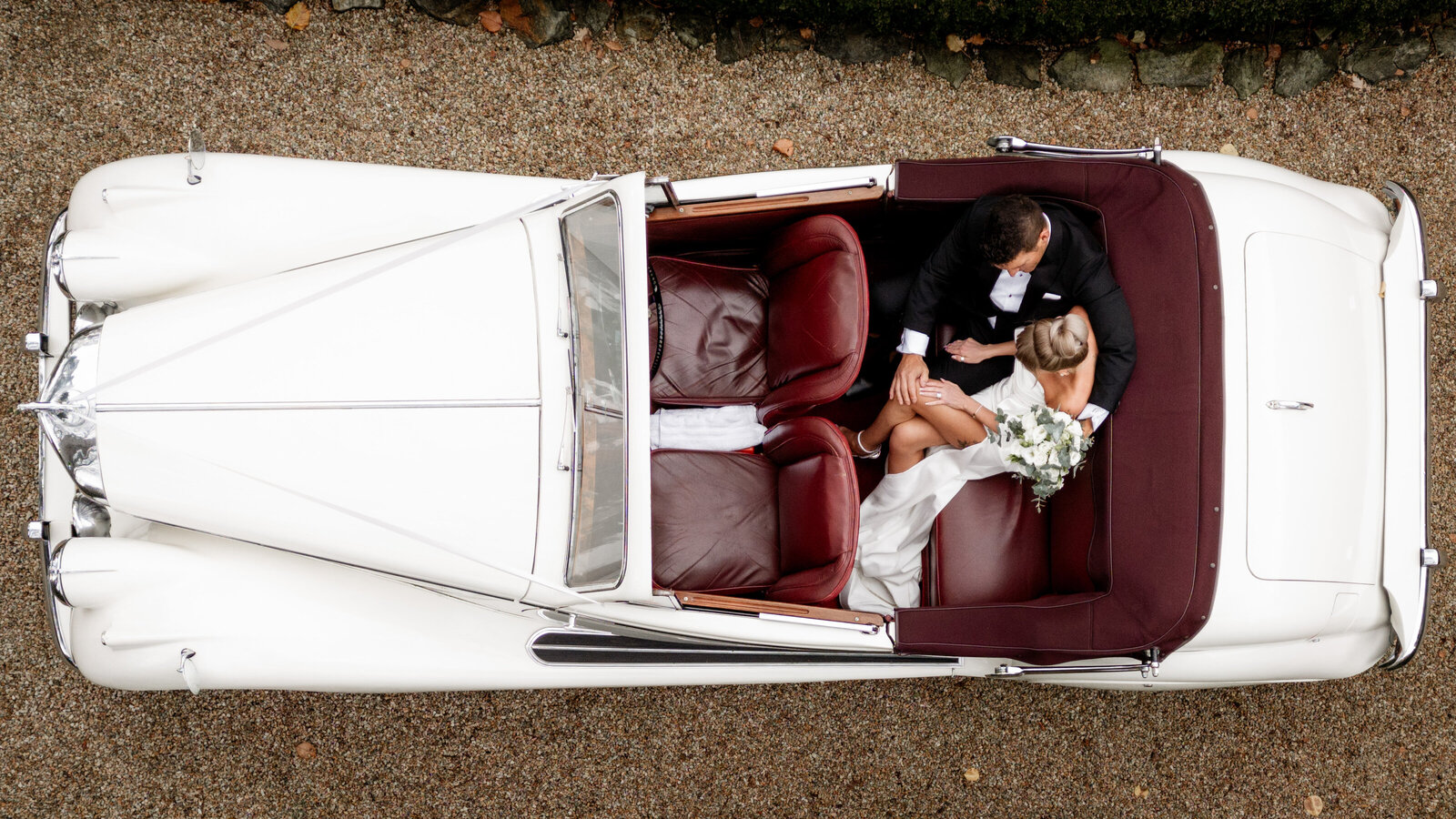Aerial view of a bride and groom sitting in a classic white convertible wedding car, captured from above to highlight the elegance and romance of their wedding day.