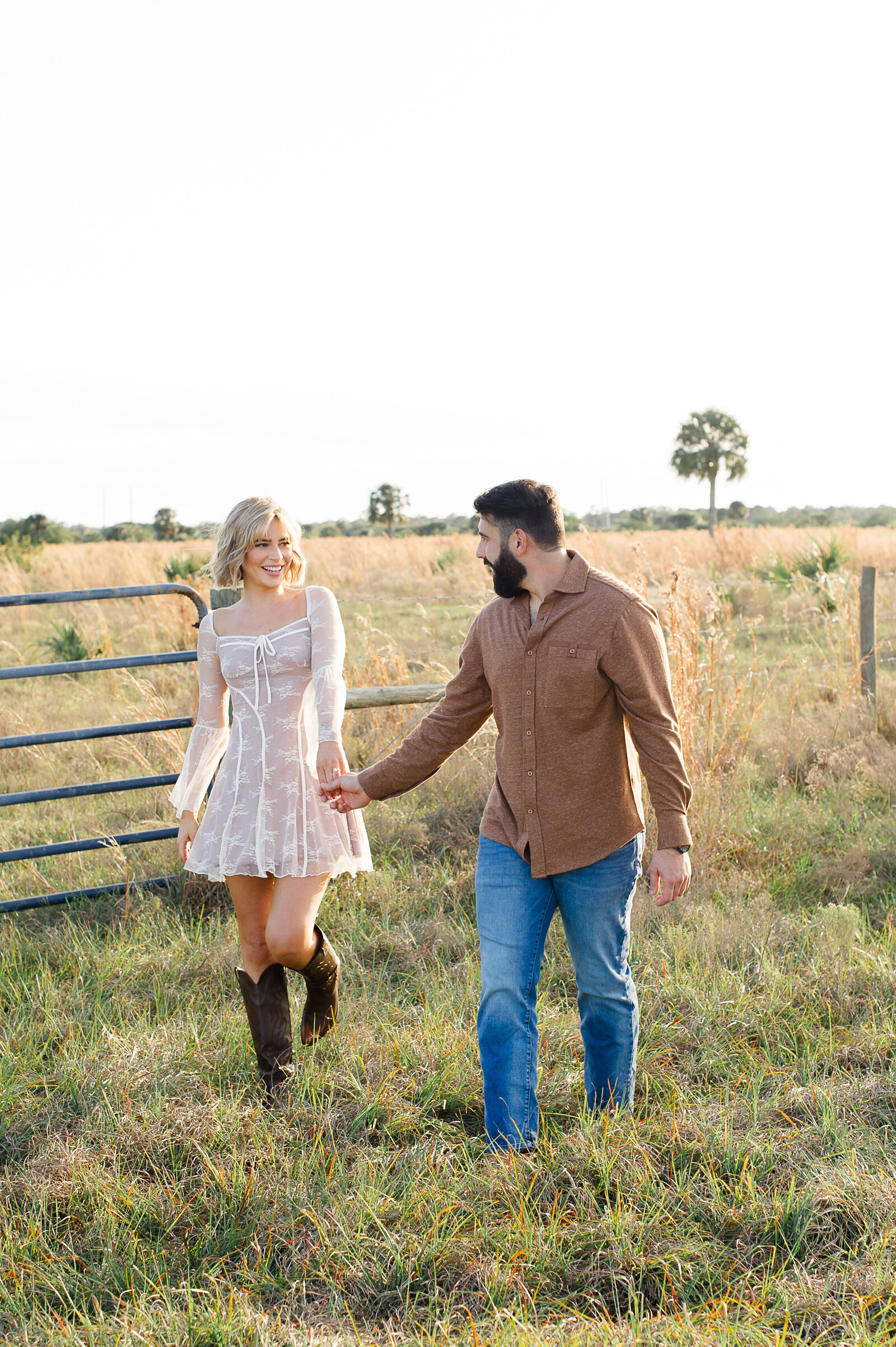 Newly engaged couple walks through a field holding hands and laughing at each other at sunset