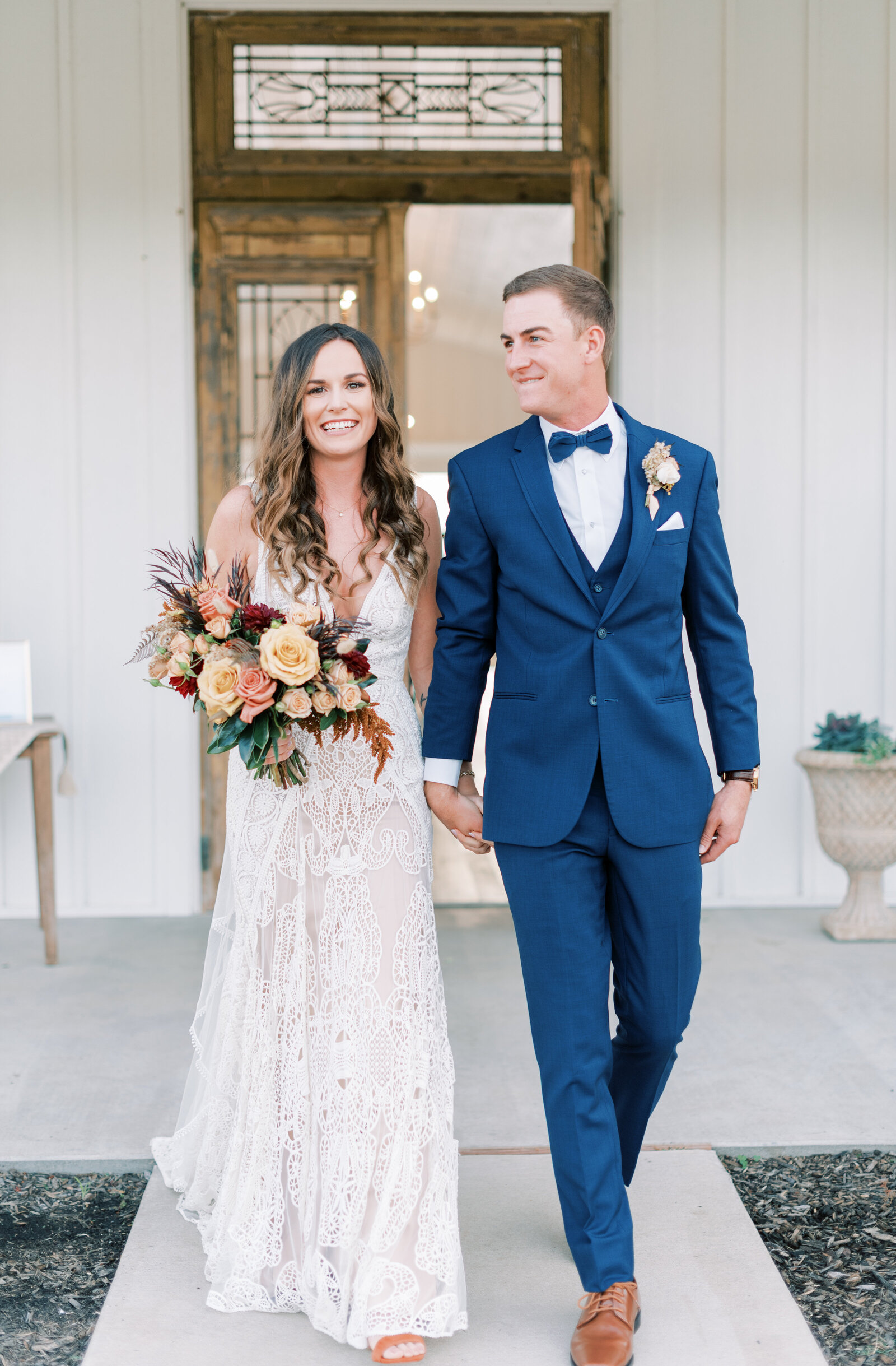 Portrait of bride and groom in a cream wedding gown and blue suit holding hands in front of  wooden door.