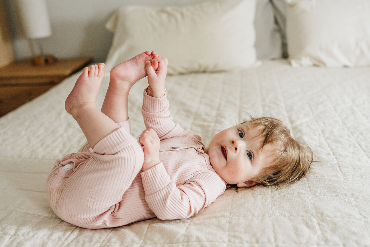 baby in pink touches toes while laying on bed