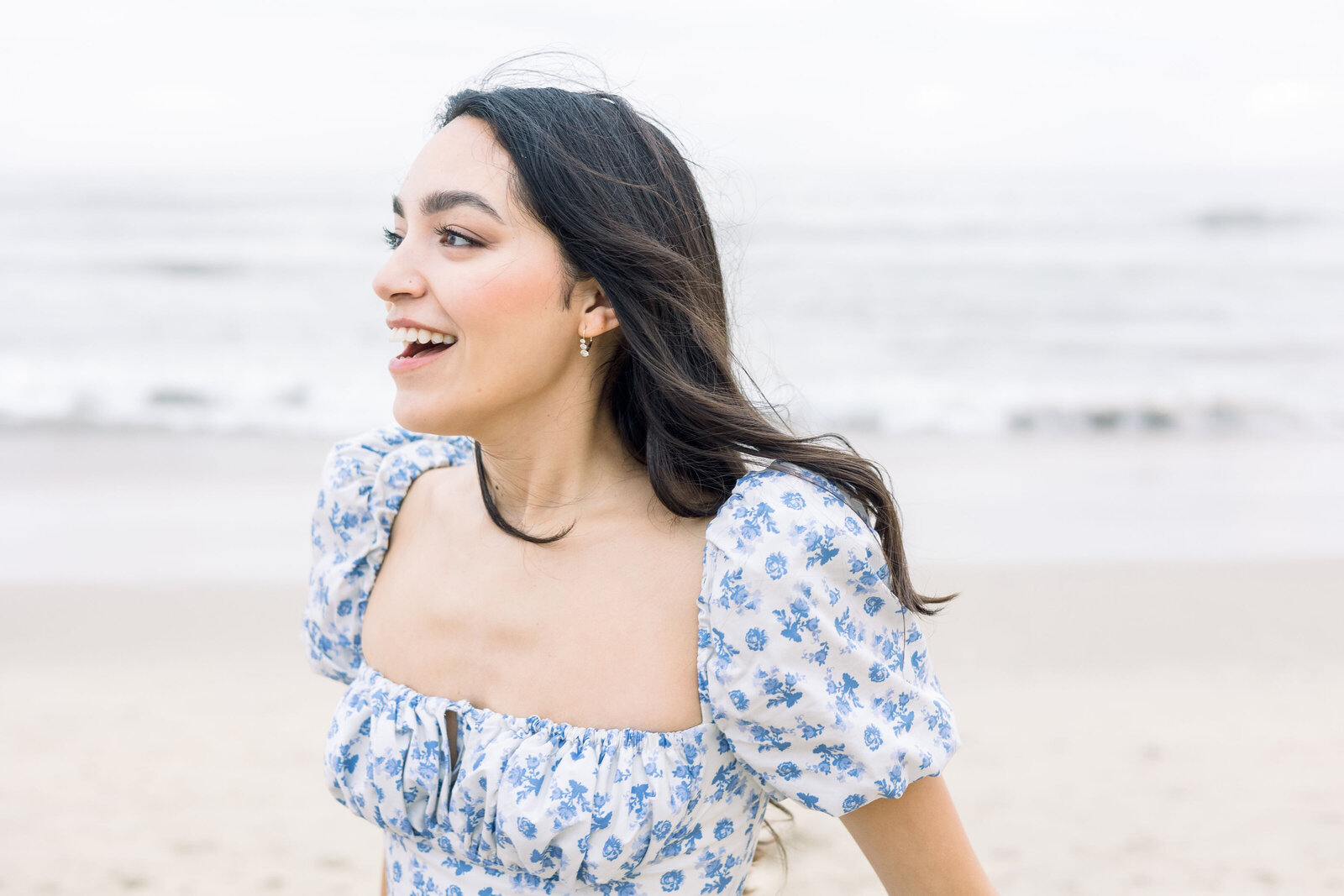 candid proposal pictures with woman with black hair in a blue and white dress smiling on the beach at half moon bay captured by wedding photographer bay area