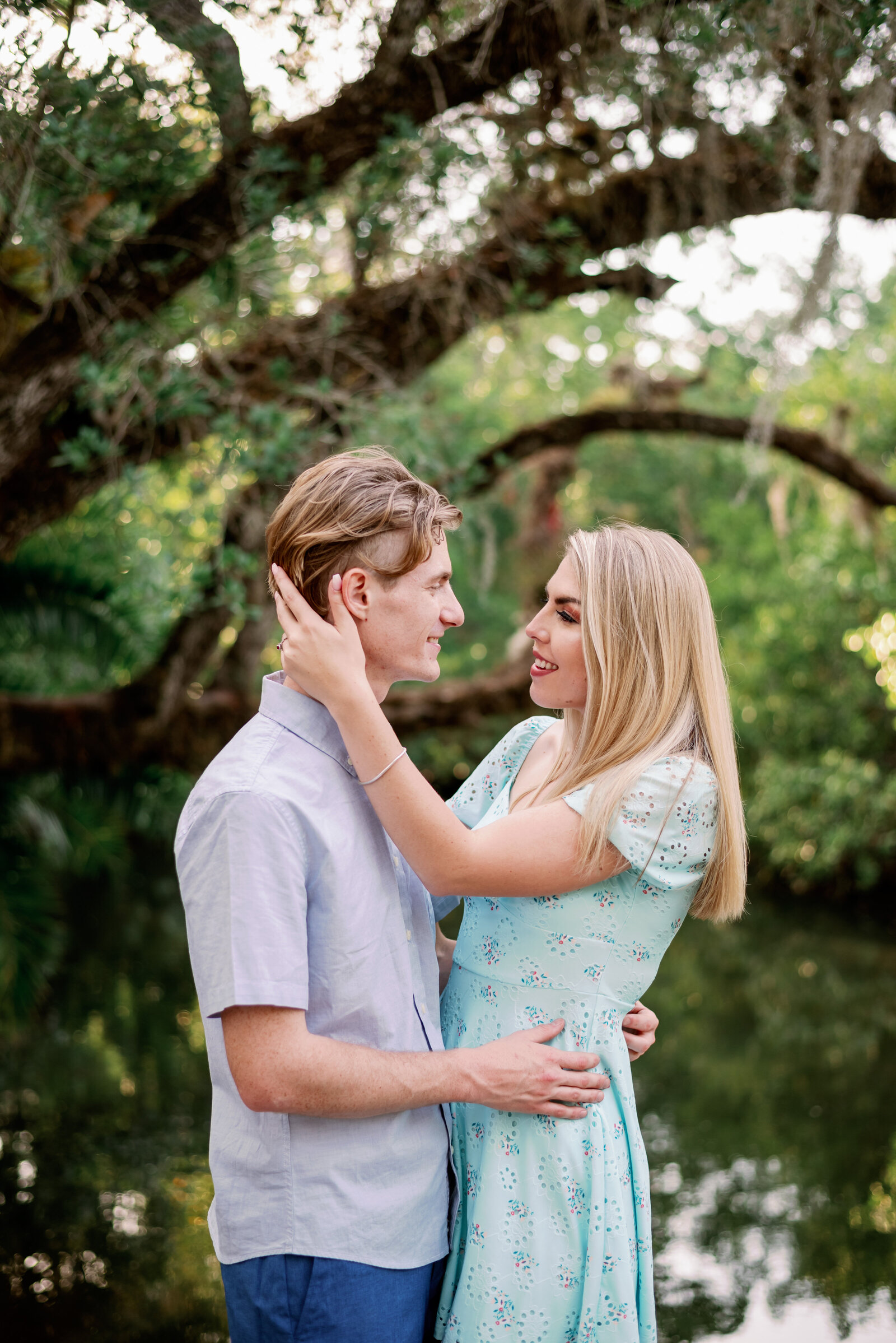engaged couple standing facing each other with water and oak trees in the background. He is holding her waist and she has her hand up toward his neck and shoulders