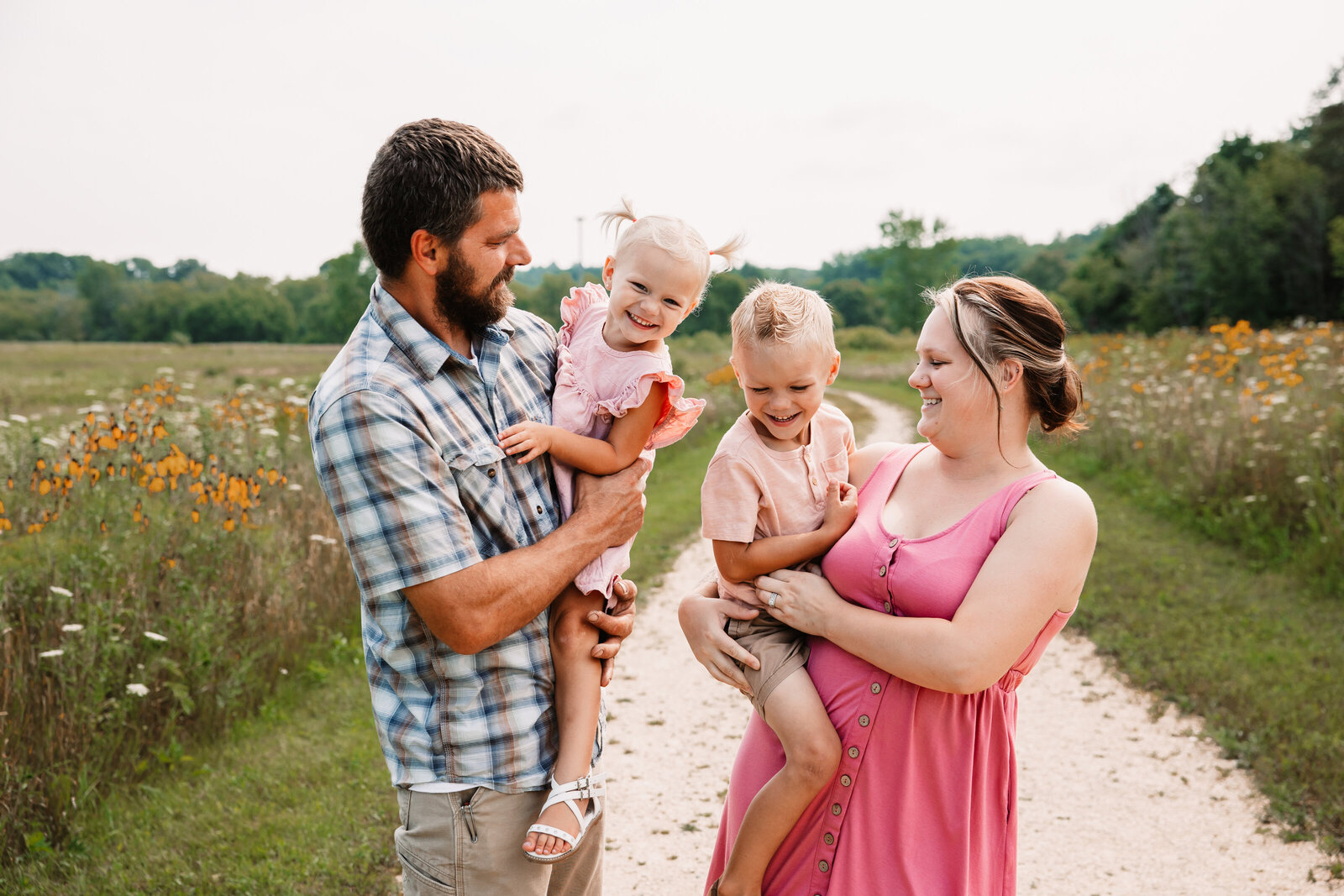 A mom and dad each carry a toddler as they walk a trail through fields of wildflowers with a Milwaukee Family Photographer