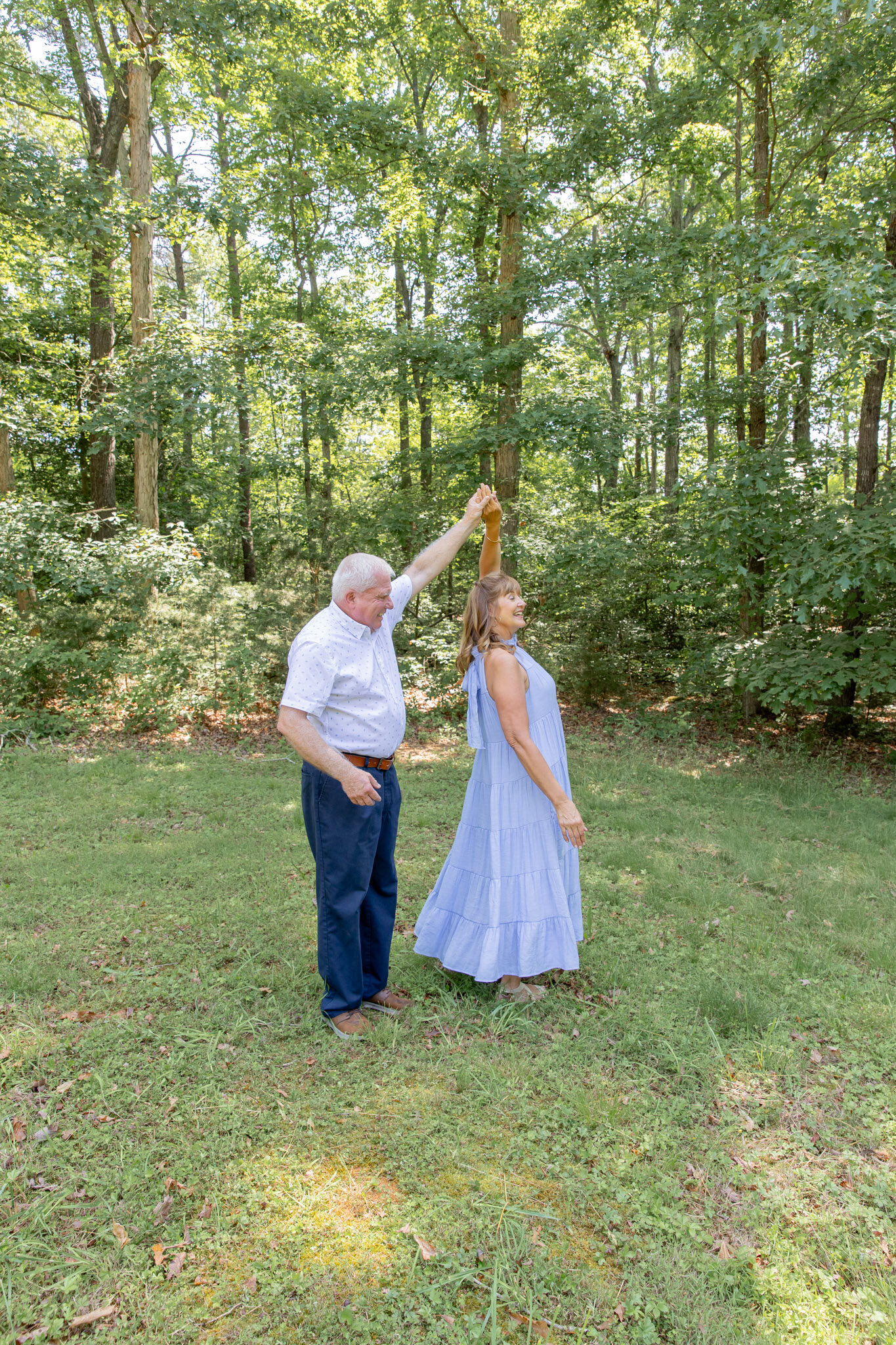 Husband twirling his wife outdoors in a field. She is wearing a blue dress and he is wearing jeans and a white short sleeved button up.
