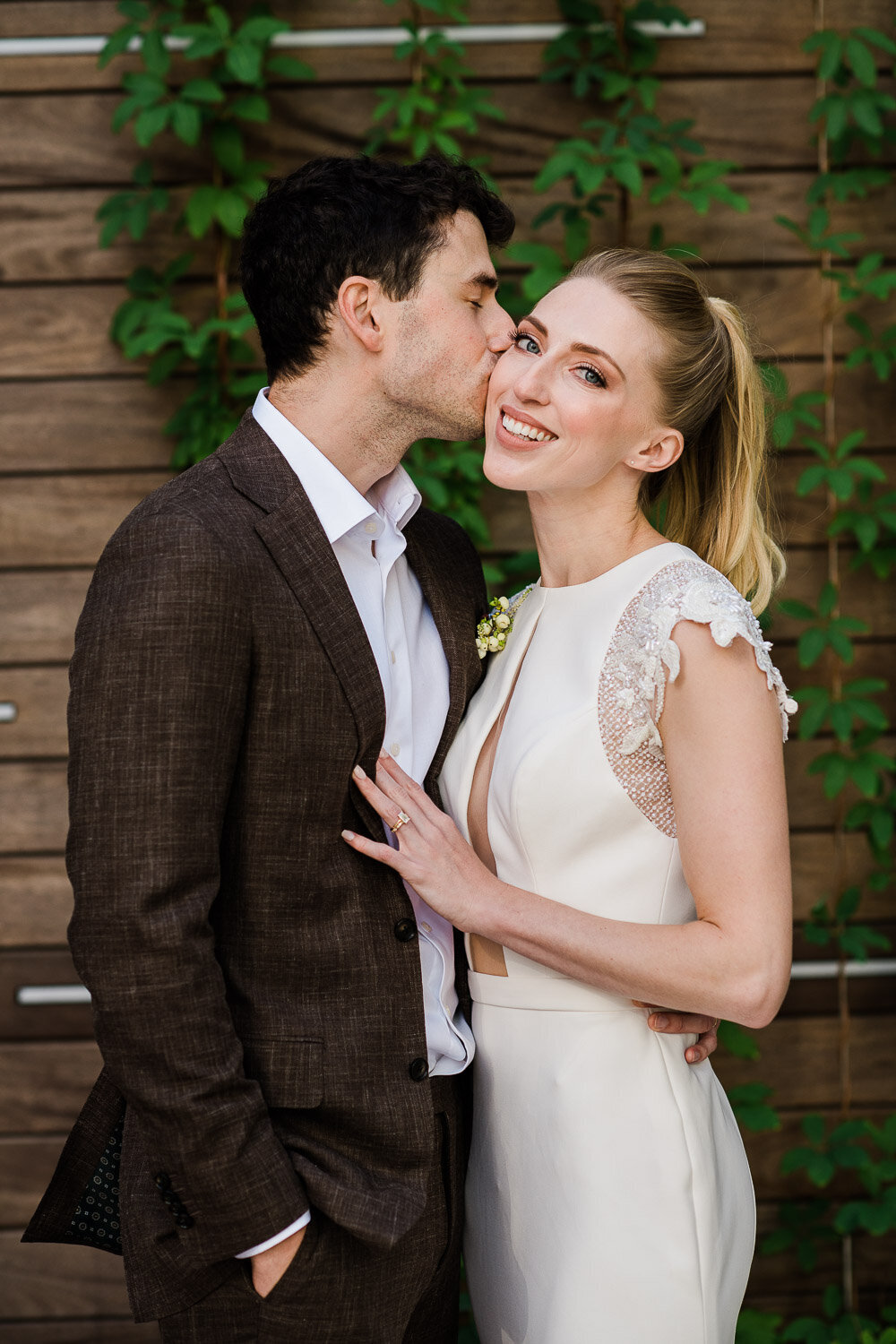 groom kiss bride cheek in front of wood and vines