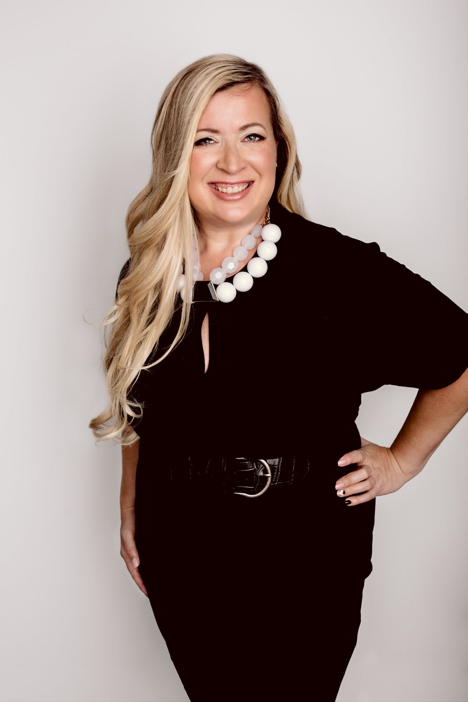 Headshot of a mid-size woman wearing a black dress and white chunky necklace against a white backdrop