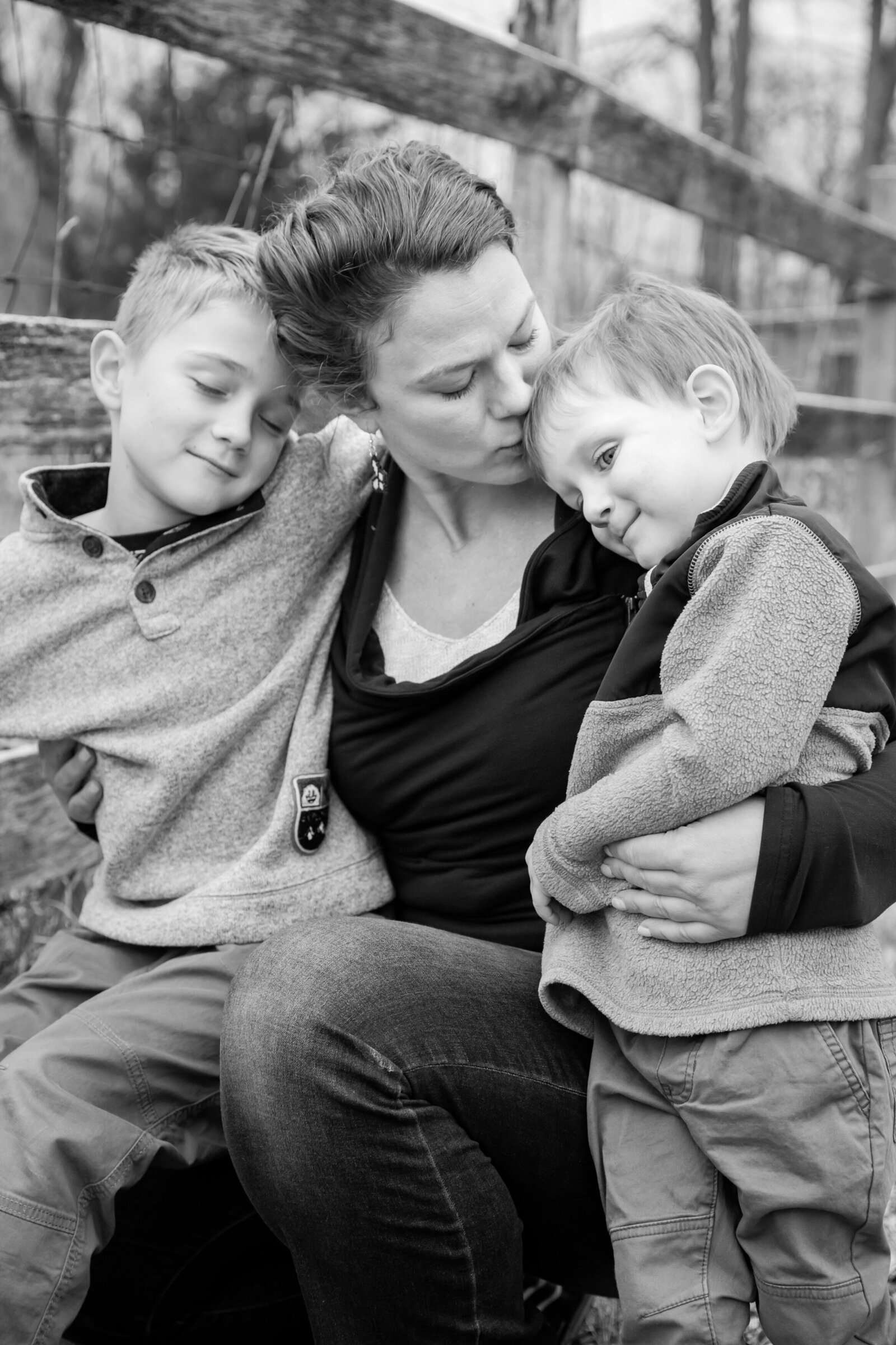 black and white photo of mother and sons snuggling with mother kissing youngest on head in pasture