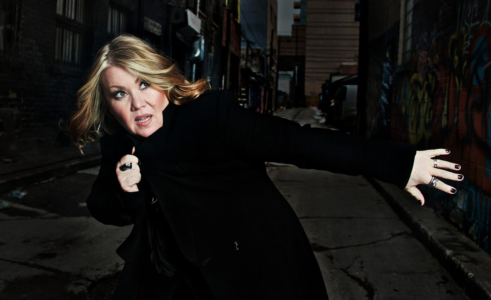 Music portrait Jann Arden wearing black overcoat with arm stretched out in alley