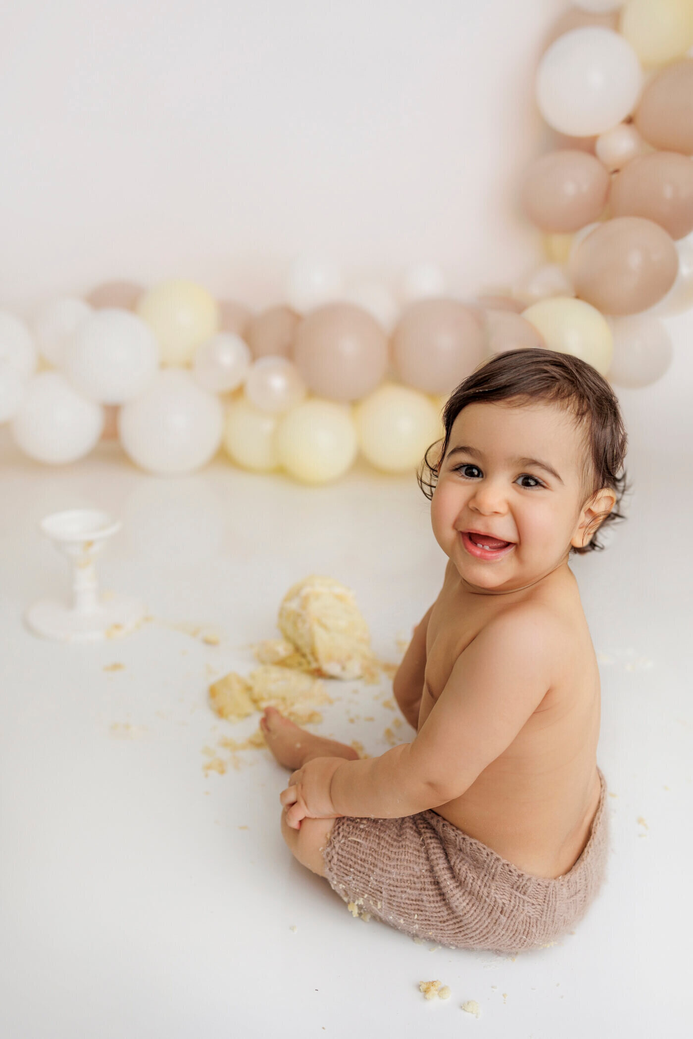 A happy baby with dark hair and eyes sits on a white floor, wearing a beige knit diaper cover, surrounded by remnants of a smashed cake. Behind the baby is a festive balloon garland in shades of white, beige, and cream. The baby looks over their shoulder, smiling joyfully at the camera.