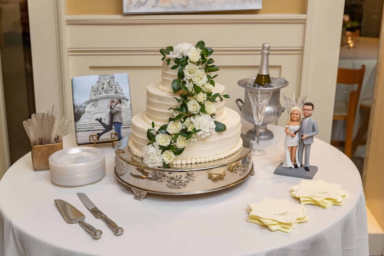 A round table displays a three-tier white wedding cake decorated with white flowers and greenery. Nearby are cake-cutting utensils, plates, napkins, a champagne bottle in an ice bucket, a photo of a couple, and a bride and groom figurine.