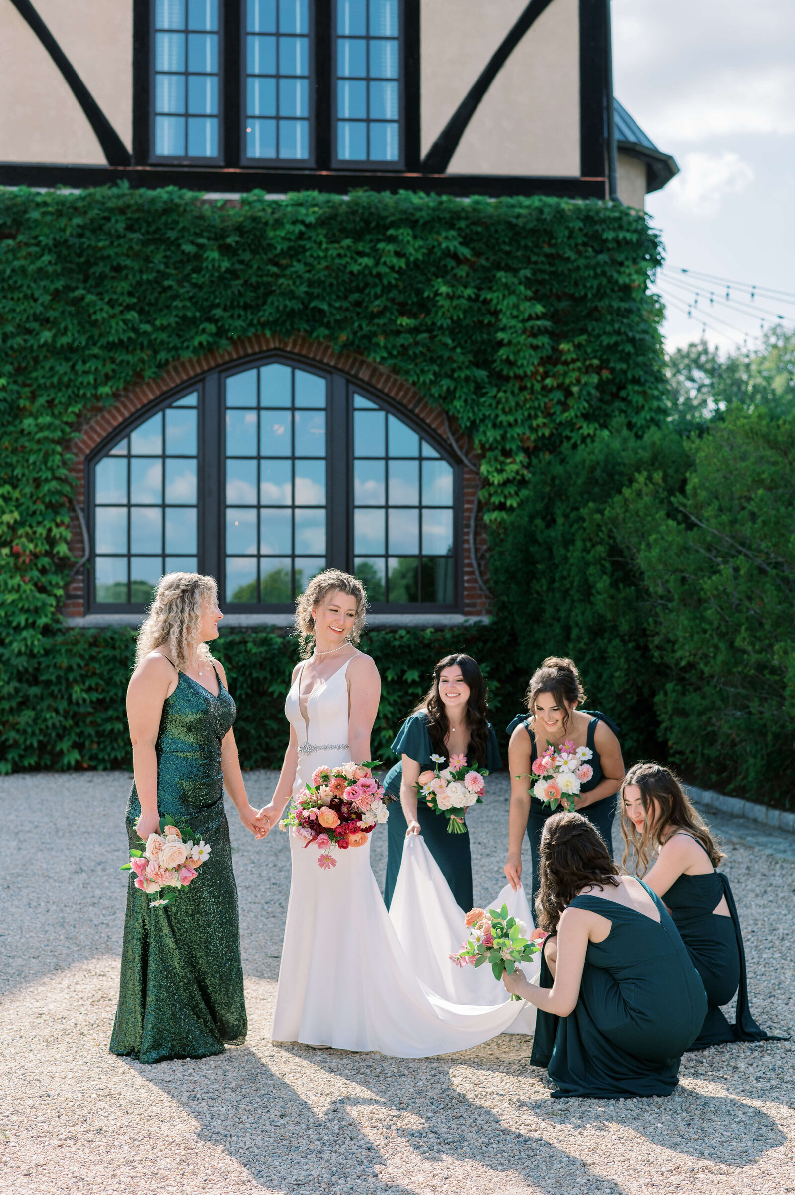 Bride in white dress smiles at her bridesmaids while they adjust her train. They are standing in front of a vine covered wall and have joyful expressions