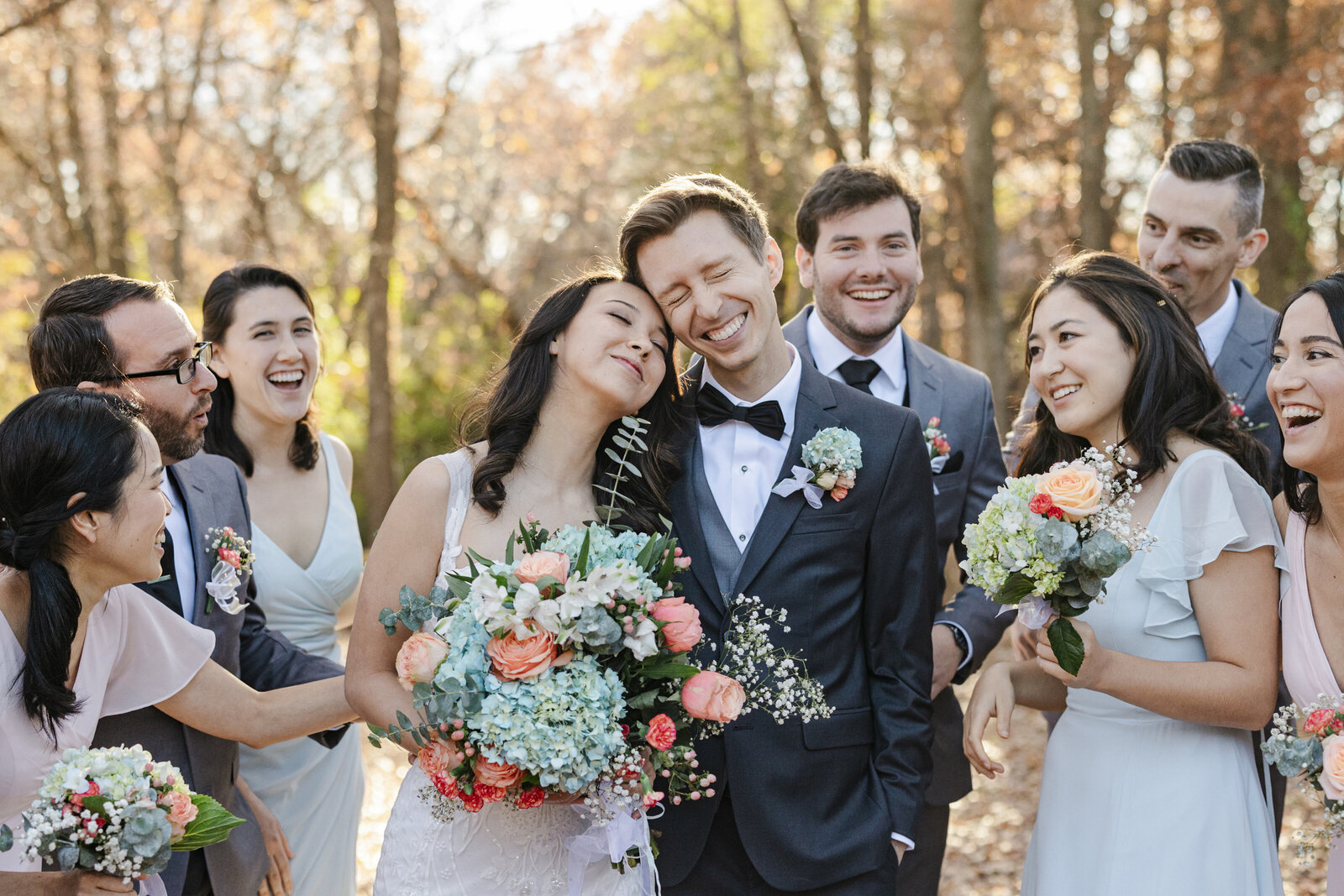 A candid portrait of a bride, groom, and their wedding party before their wedding ceremony in DFW, Texas. The bride is in the center on the left and is leaning her face against the groom's face while holding a large, colorful bouquet. She is wearing a sleeveless, white, highly detailed white dress. The groom is right next to her and is smiling widely with closed eyes. He is wearing a three piece suit with a bowtie and boutonniere. The groomsmen are all wearing a grey suits with ties and boutonnieres while the bridesmaids are wearing dresses in shades of light pink and light blue while holding small, colorful bouquets. They are all backed by a large row of fall trees.