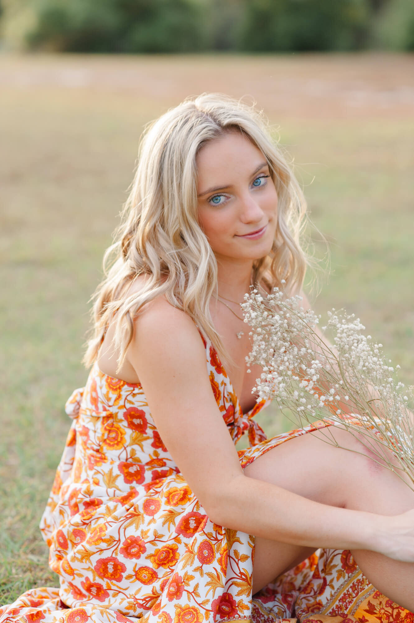 Blonde senior wearing gorgeous orange floral dress holds babys breath and sits in the grass smiling at the camera