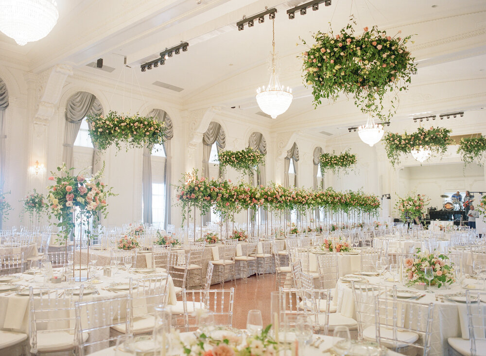 A large ballroom full of a wedding reception and large floral arrangements on every table as well as large installations hanging from the ceiling.