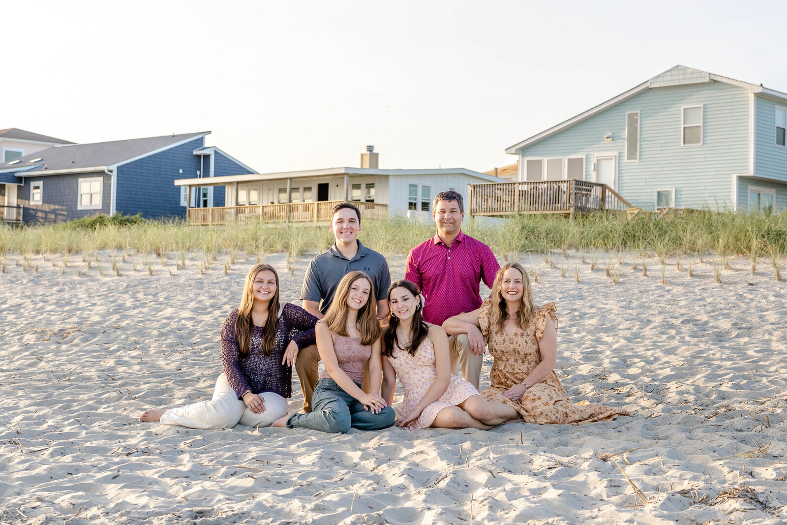  A family sits on the sandy beach in front of colorful coastal homes. Dressed in casual summer attire, they enjoy a sunny day, ideal for professional family photography in NC, showcasing relaxed and vibrant beach gatherings.
