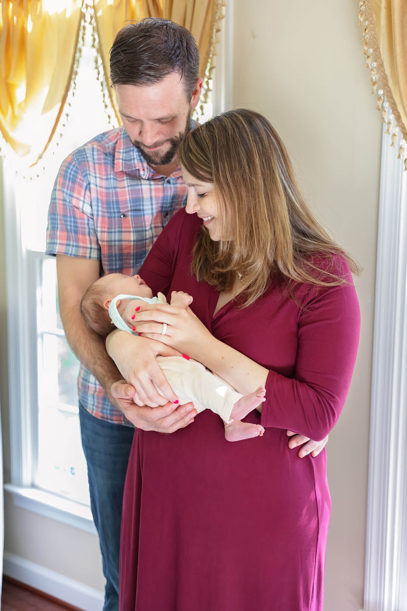 A mom and dad standing with their newborn daughter in front of a window.