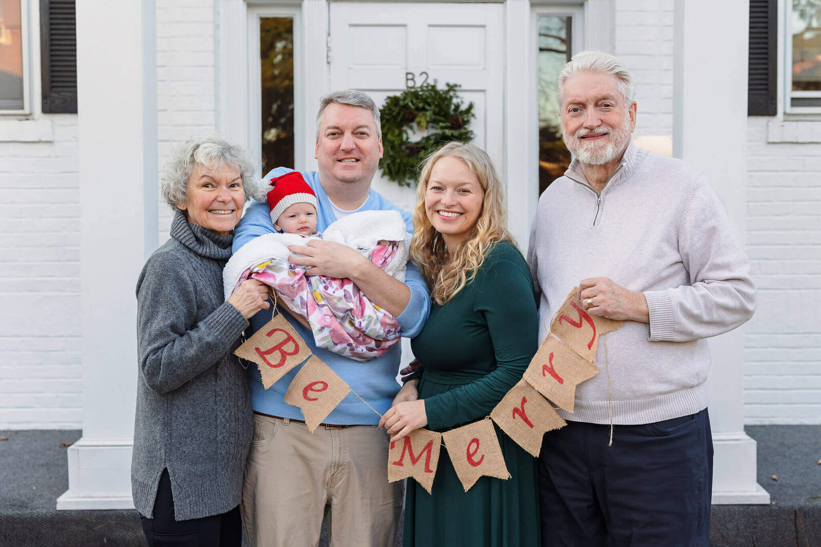 A close-up of an extended family holding a Be Merry sign in front of a white building.
