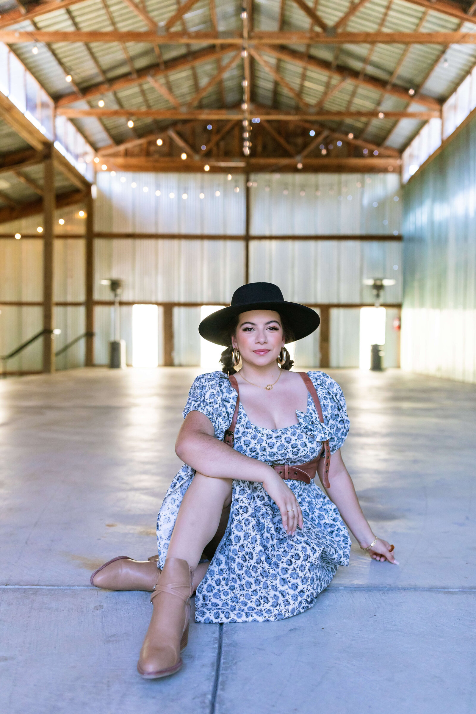 dark haired girl wearing black hat, brown belt, and blue floral dress with bubble braids in a warehouse