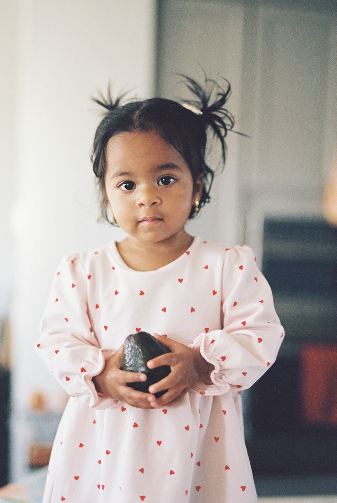 "A tender portrait of a young girl with pigtails, wearing a heart-patterned dress, holding an avocado with an innocent and curious expression, captured in soft natural light.