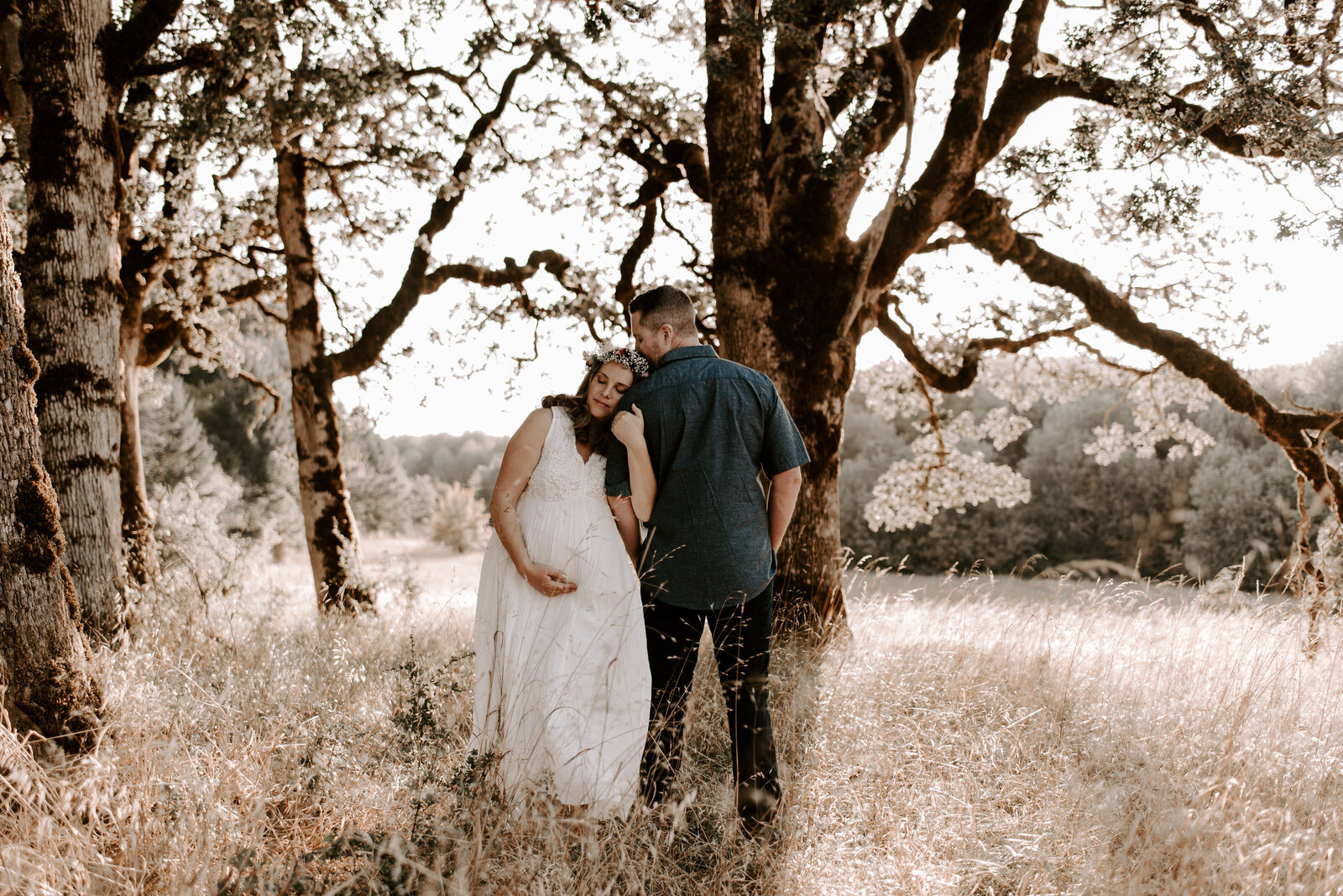husband and wife holding pregnant belly in field with oak trees