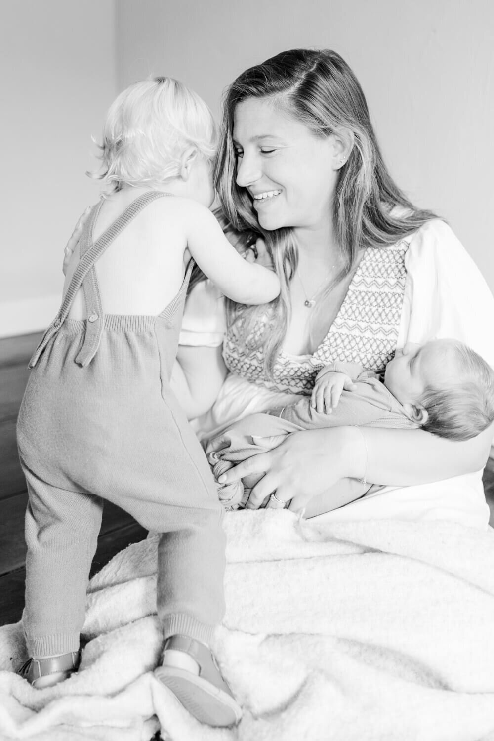 Black and white image of mom playing with toddler while holding newborn