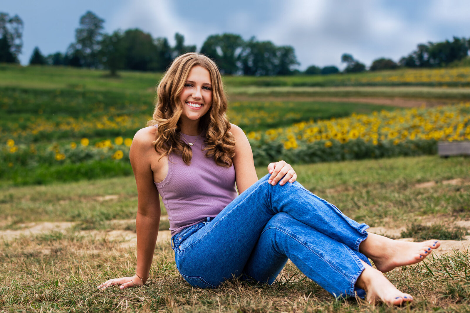 This girl sits on the ledge of a field wearing a purple shirt and blue jeans her bare feet are crossed in front of her and she smiles at the camera for senior portraits.