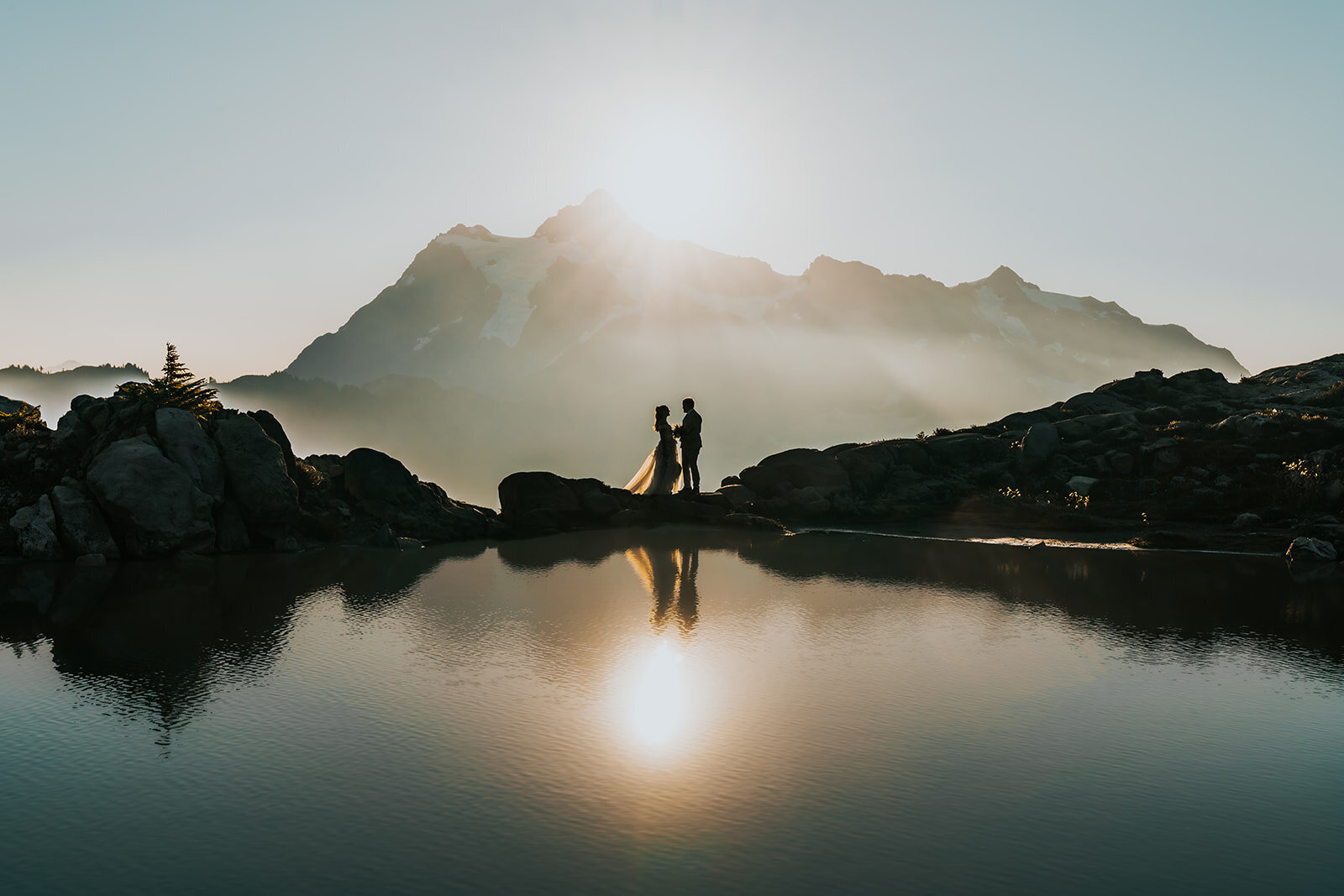 a panoramic image of sweeping mountains at sunrise. A couple, in their wedding attire embraces on a peak in front of a reflective tarn as their adventure elopement photographers capture the moment. 