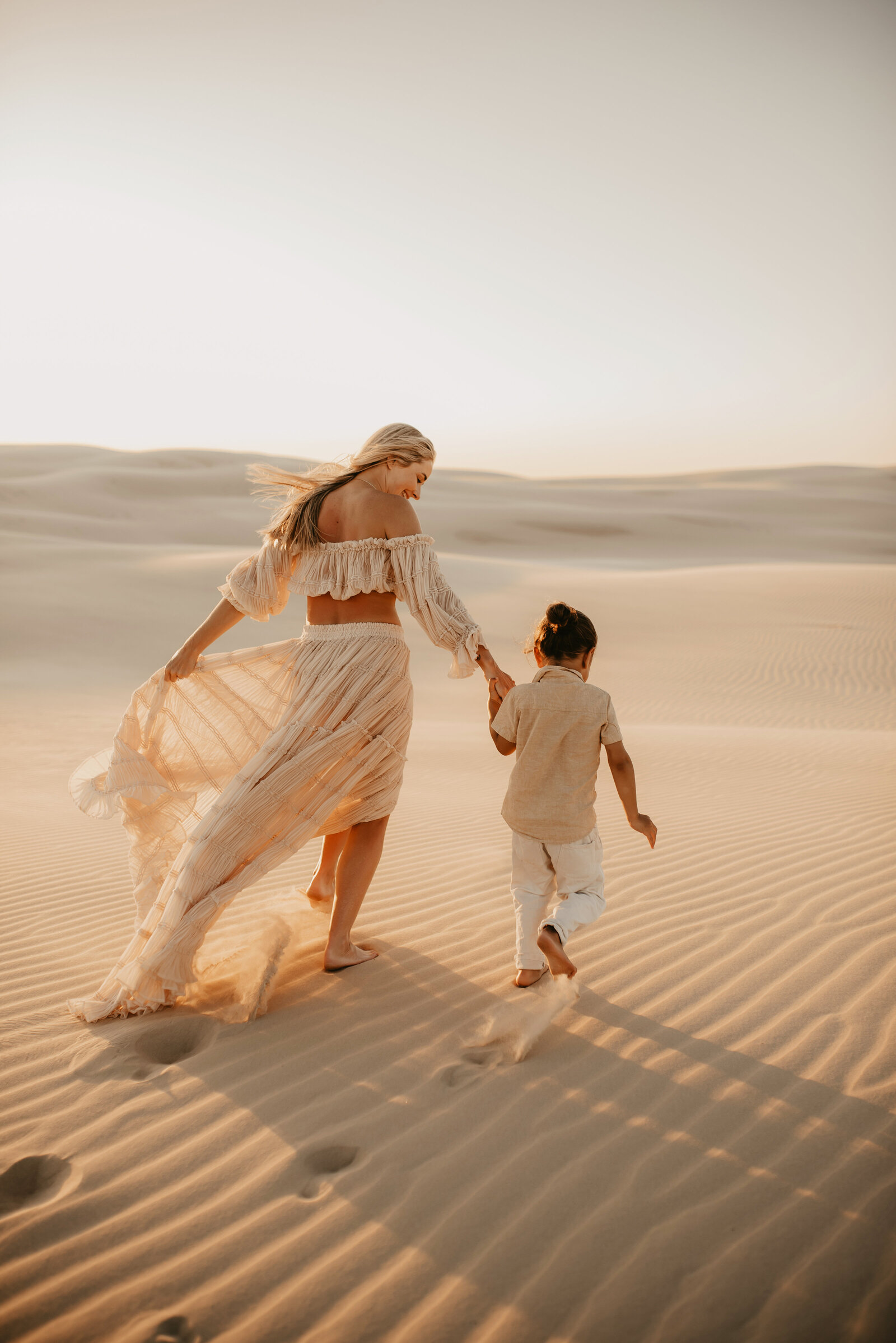 A woman in a flowing dress holds hands with a child as they walk across sand dunes at sunset.