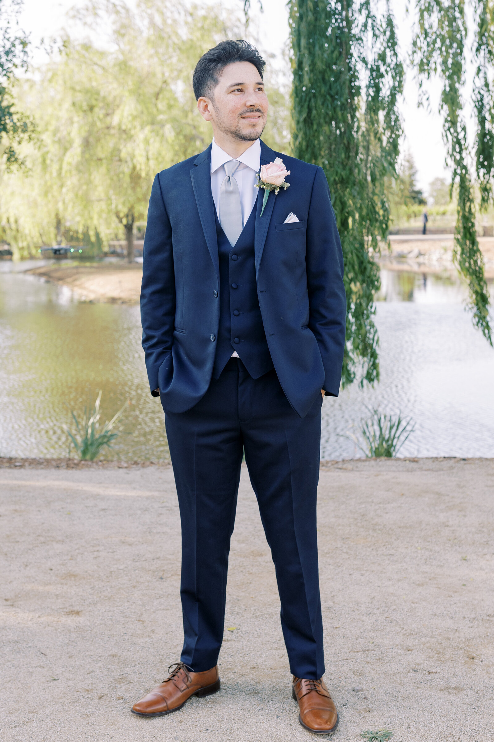 sacramento wedding photographer photographs groom's portrait with groom in a blue suit standing in front of a lake at Wolfe Heights wedding venue with weeping willows in the distance