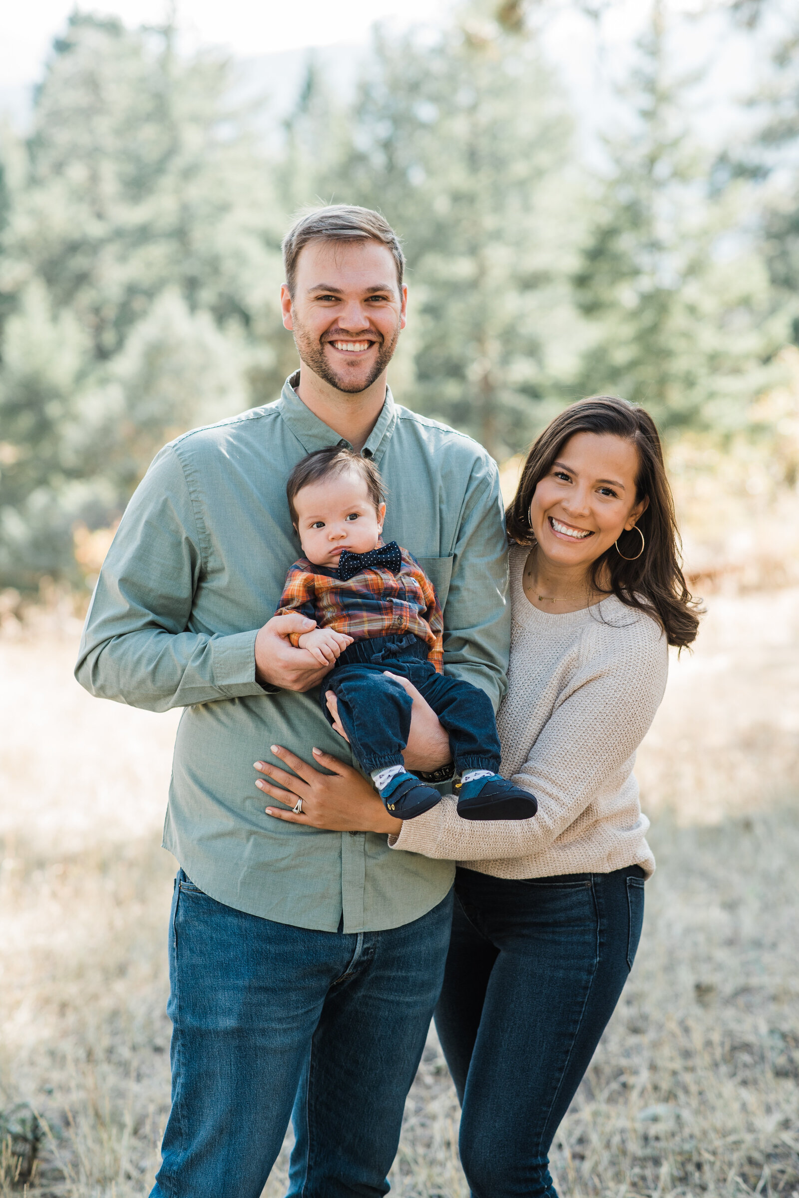 family outdoor photos with dad holding a newborn baby boy while wearing a green button down as the mom leans in and hugs her family and smiles captured by denver family photographers