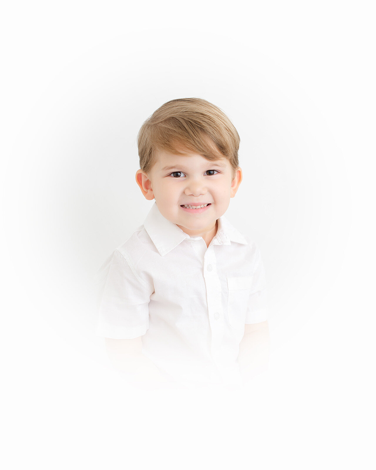 A young child with light brown hair and a light complexion smiles at the camera, captured by a talented photographer in a white short-sleeved shirt against a plain white background. This image is destined to become a timeless heirloom.