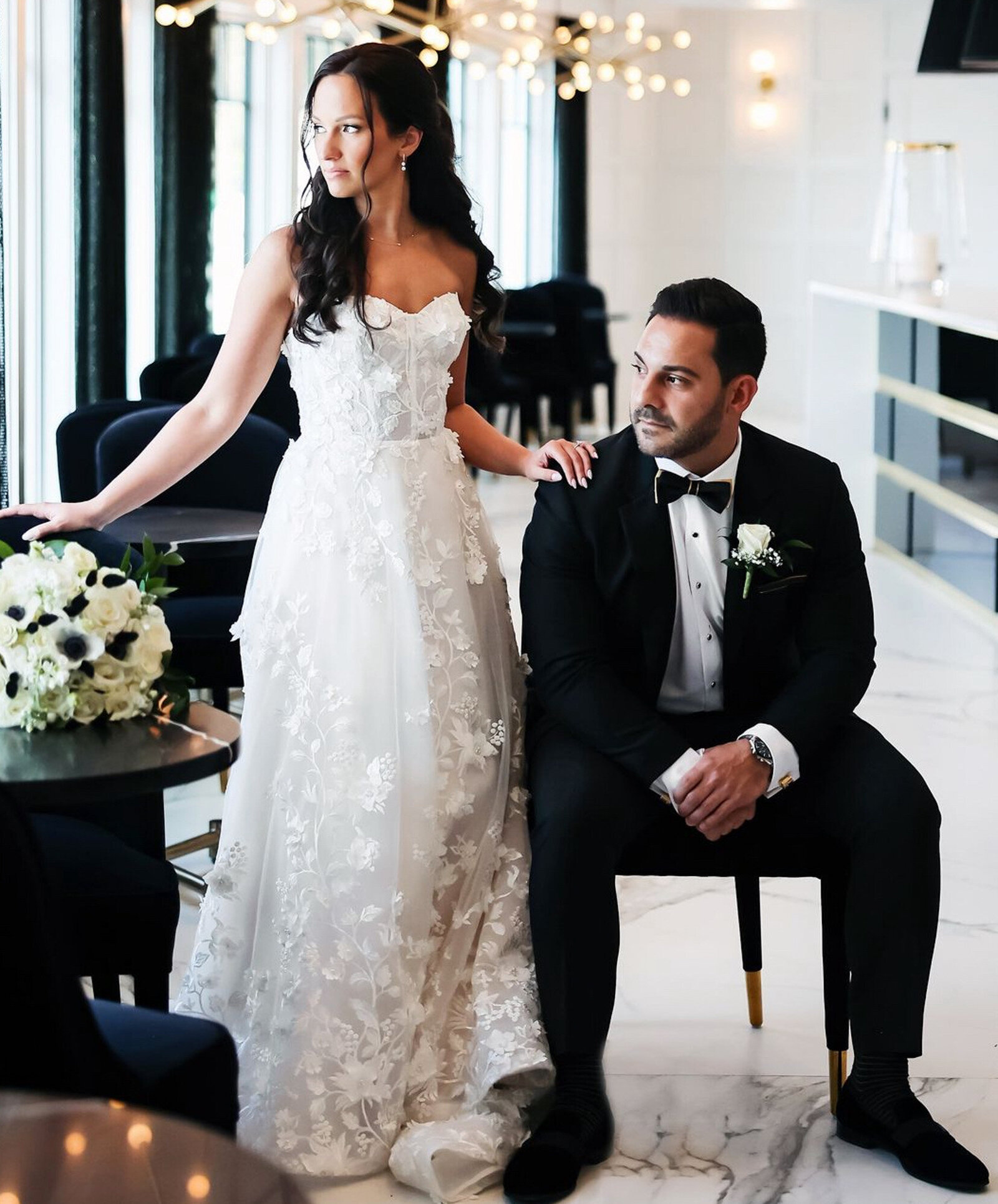 bride, in a white floral lace gown, stands next to a groom seated on a chair in a black tuxedo with a bow tie.\