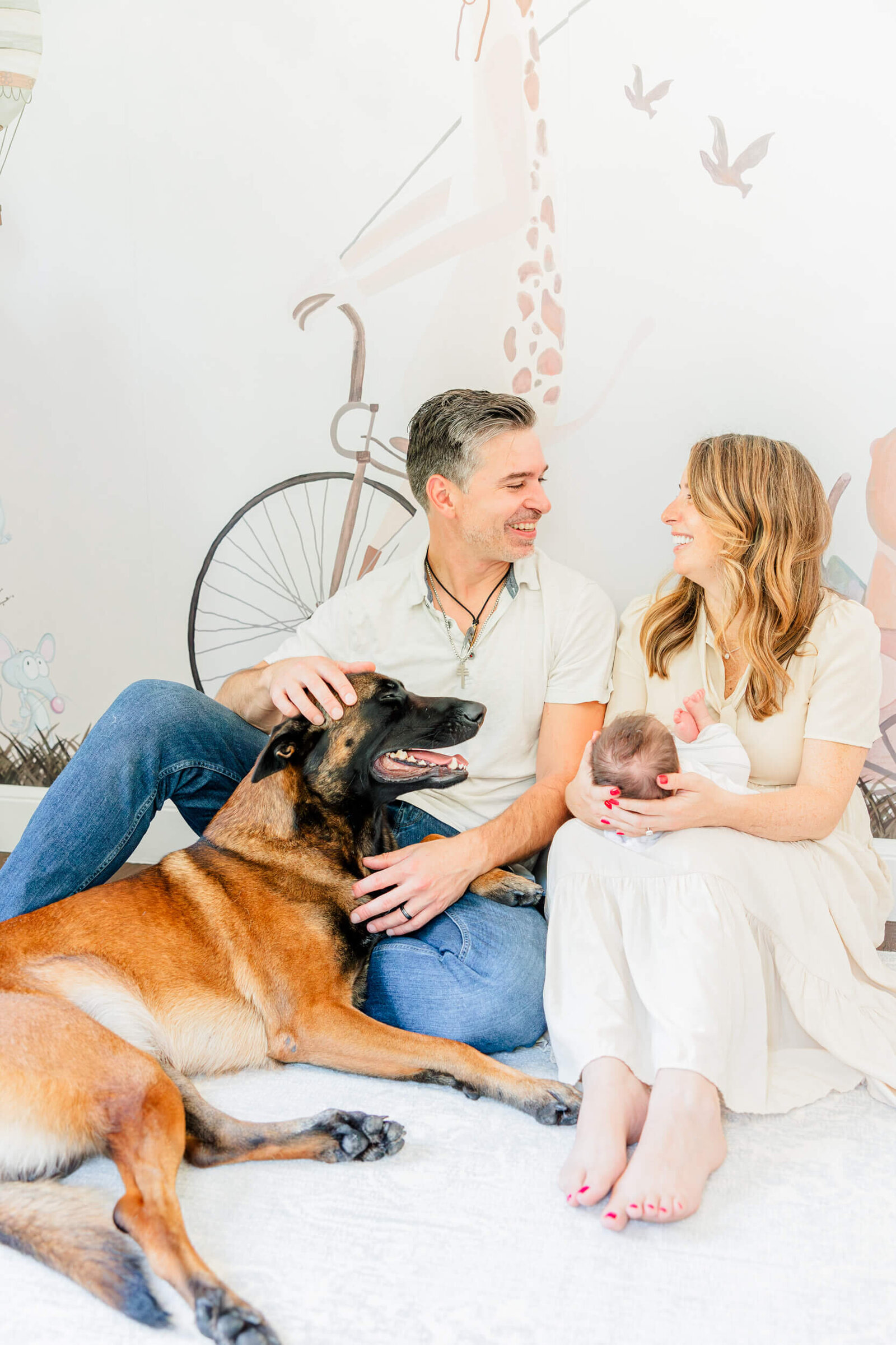 Dad pets a Belgian Malinois and laughs with mom, who is holding their baby