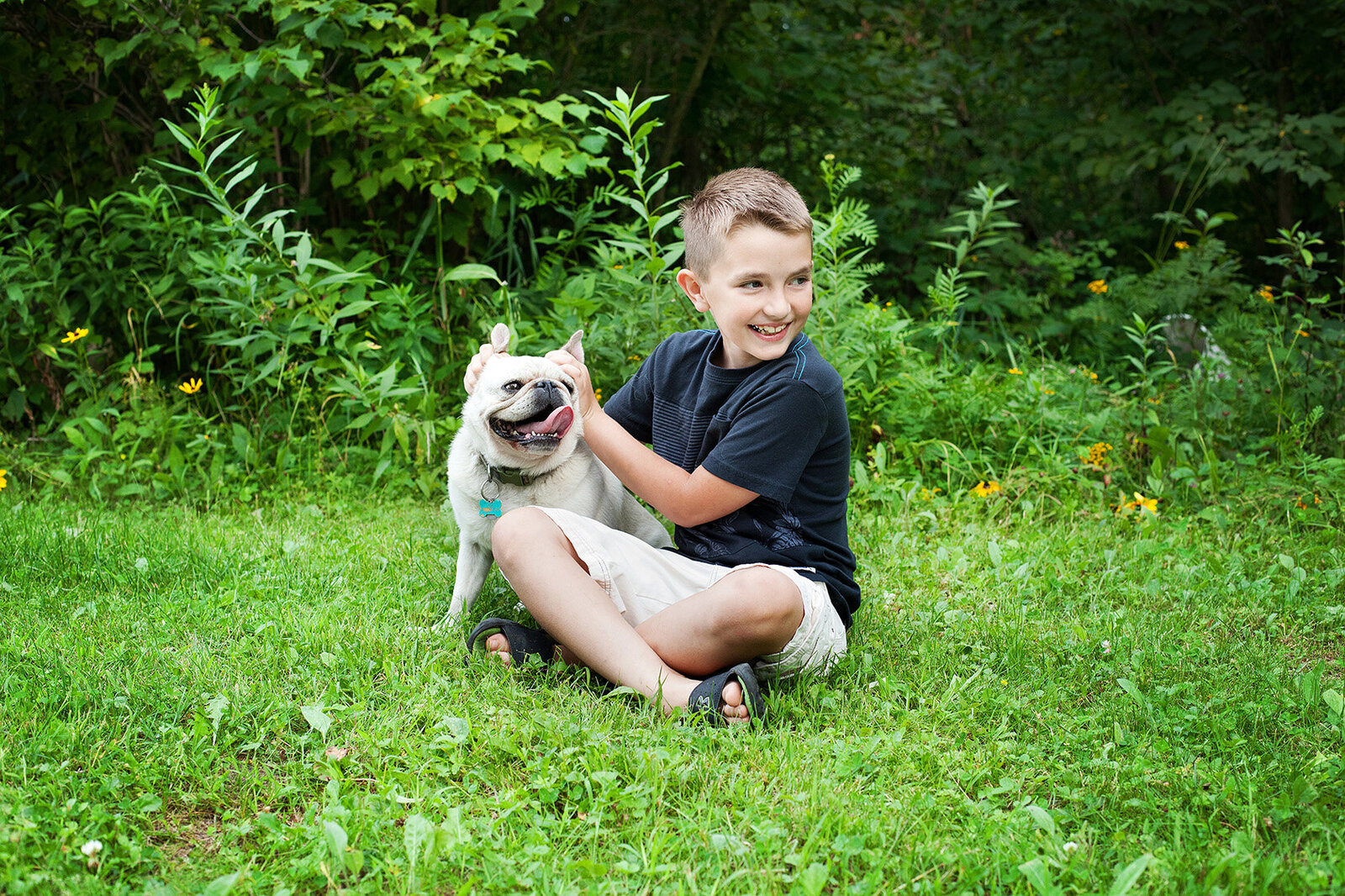 A young pre-teen boy sitting on the grass holding his dogs ears up and laughing.