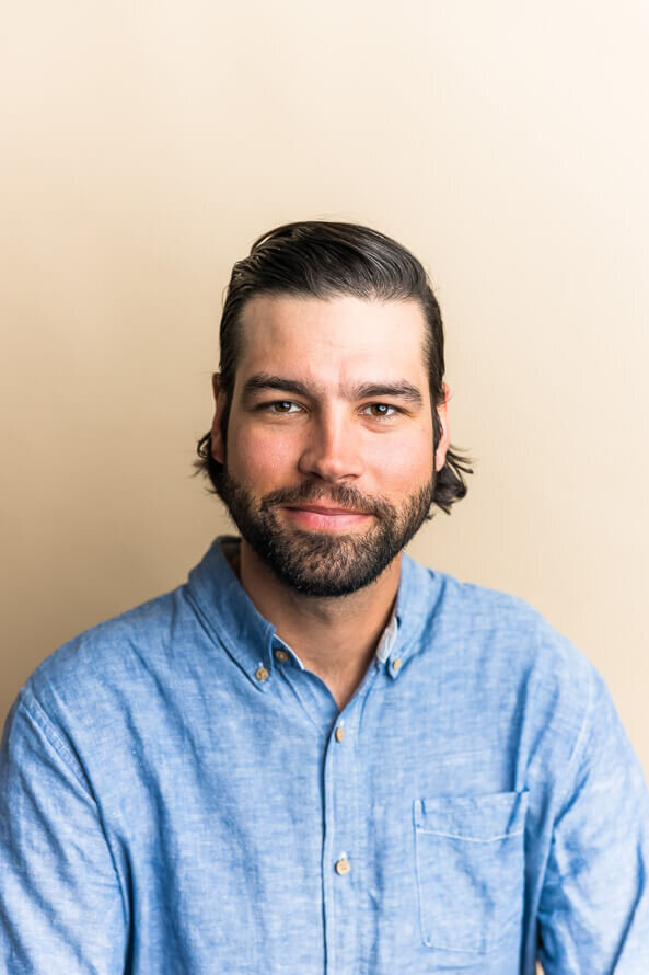 Man with beard poses for his headshot on a tan backdrop.