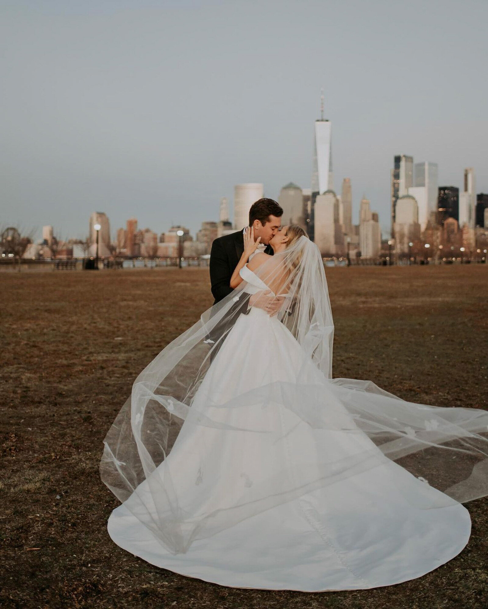 bride wears a long white dress with a flowing veil, and the groom is dressed in a black suit shares a kiss in an open field with a cityscape, including a tall, prominent building, visible in the background