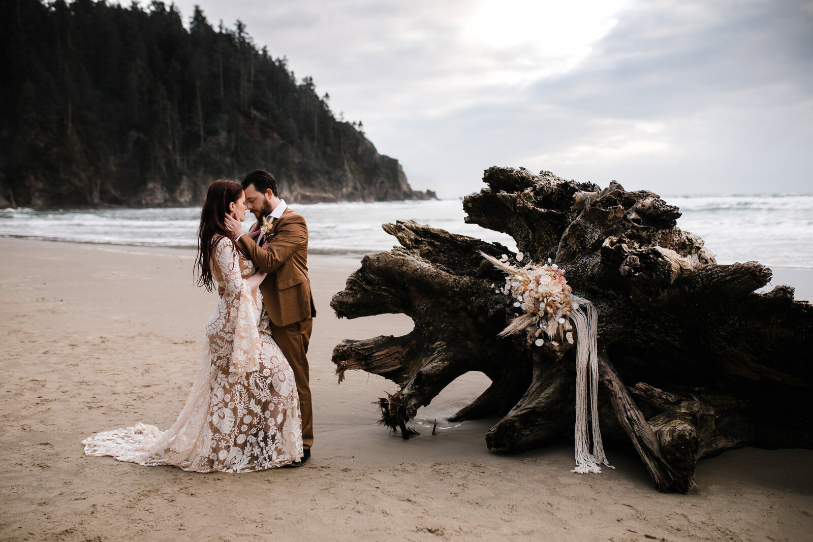 A moody beachside elopement session framed by driftwood and rugged coastal scenery, perfect for adventurous souls.