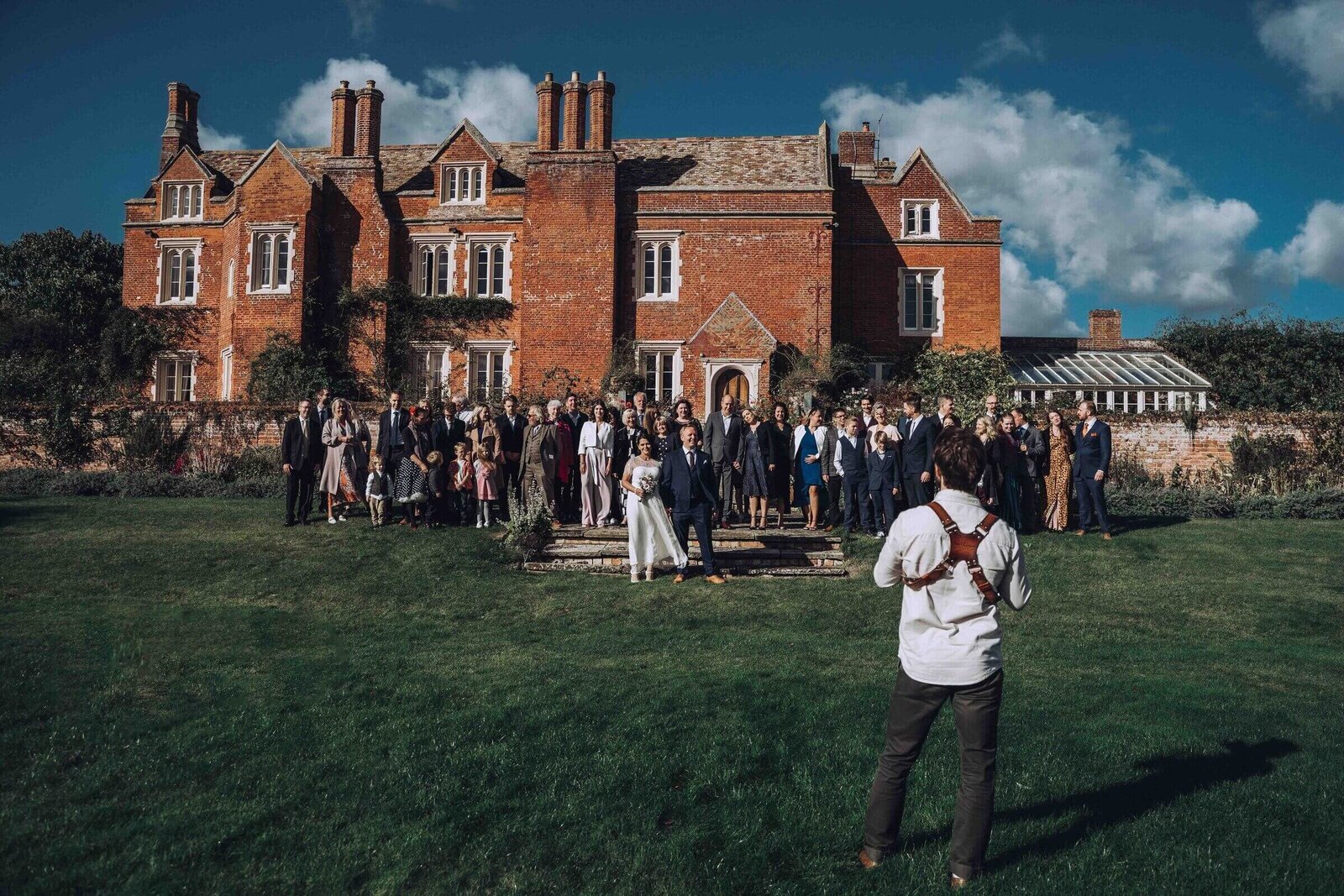 Tom Keenan photographing a large group of wedding guests arranged in front of a stately building, capturing formal group portraits.