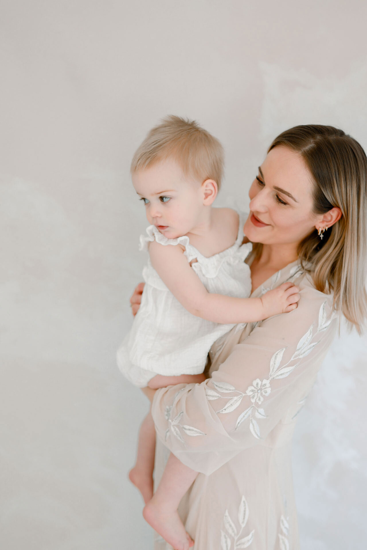 Bei der Babyfotografin in Bielefeld während einem Baby Fotoshooting hält Mama, die ein zartes weißes Kleid an hat ihr Baby auf dem Arm, das nach etwas Ausschau hält.