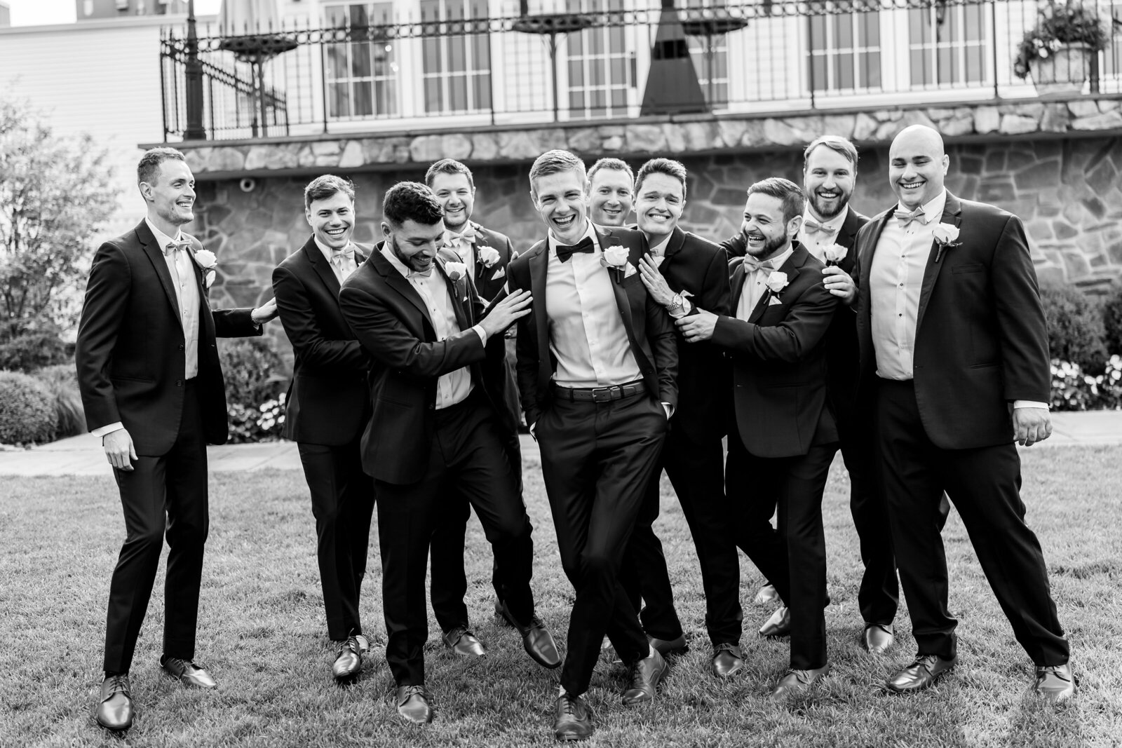 A group of groomsmen in suits and bow ties, gathered outdoors, joyfully surrounding the groom.