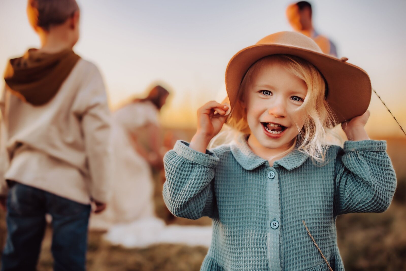 A little girl holding her hat and smiling into the camera with her family blurred in the background behind her during their family photoshoot captured by Infinite Productions