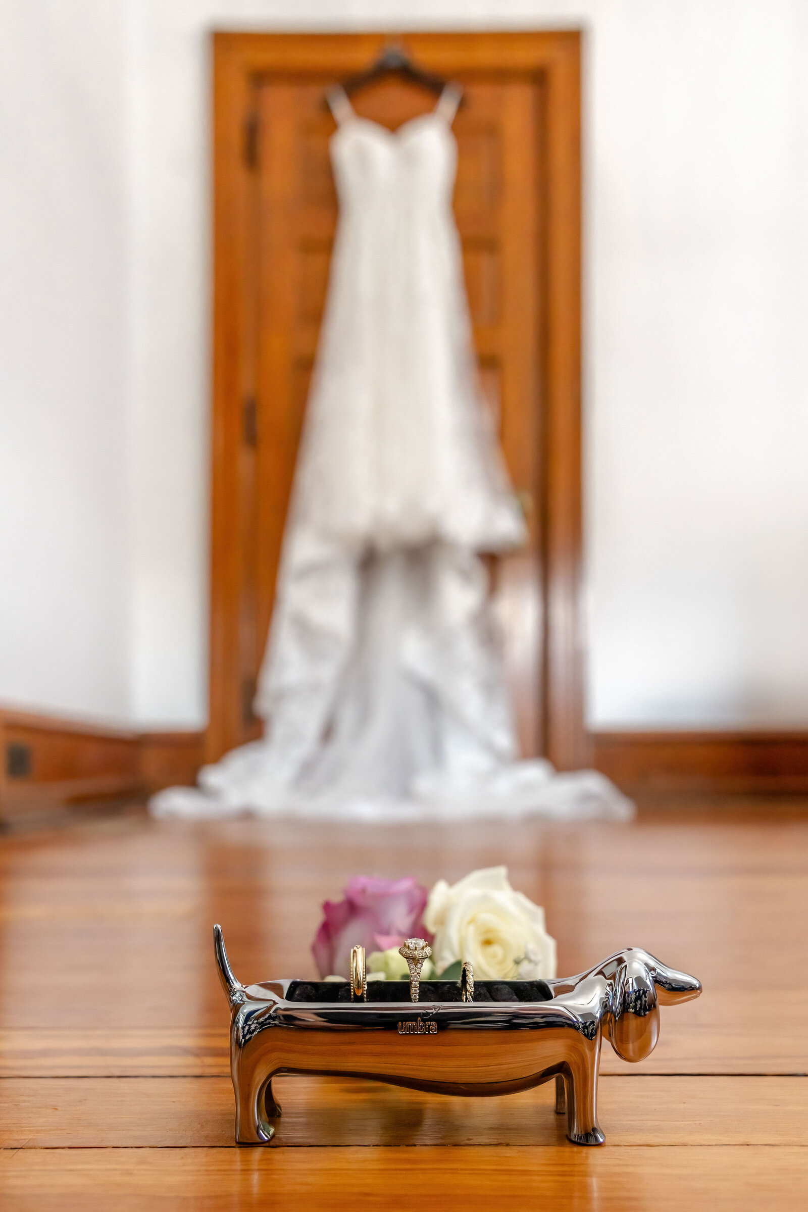 Wedding rings displayed on a silver dachshund ring holder with a blurred wedding dress and floral accents in the background, captured during a Wilmington, NC wedding, blending creativity and elegant details.