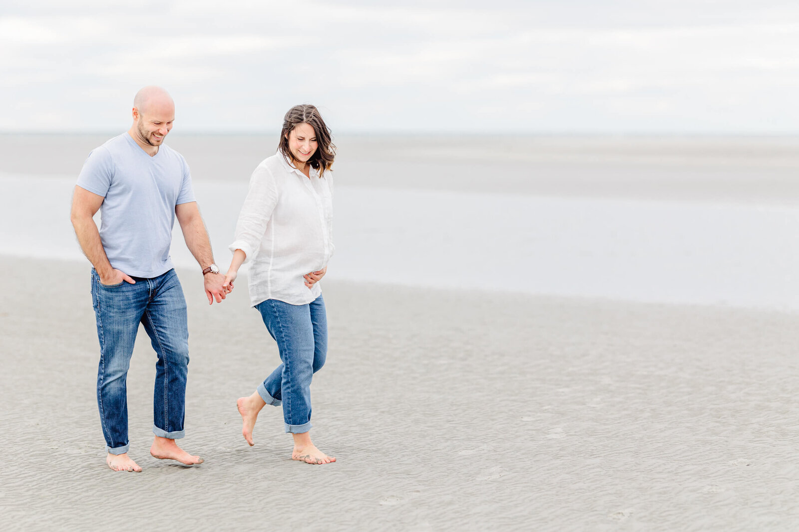 Pregnant woman and her husband walk along the ocean on a cloudy day at Boston's northshore