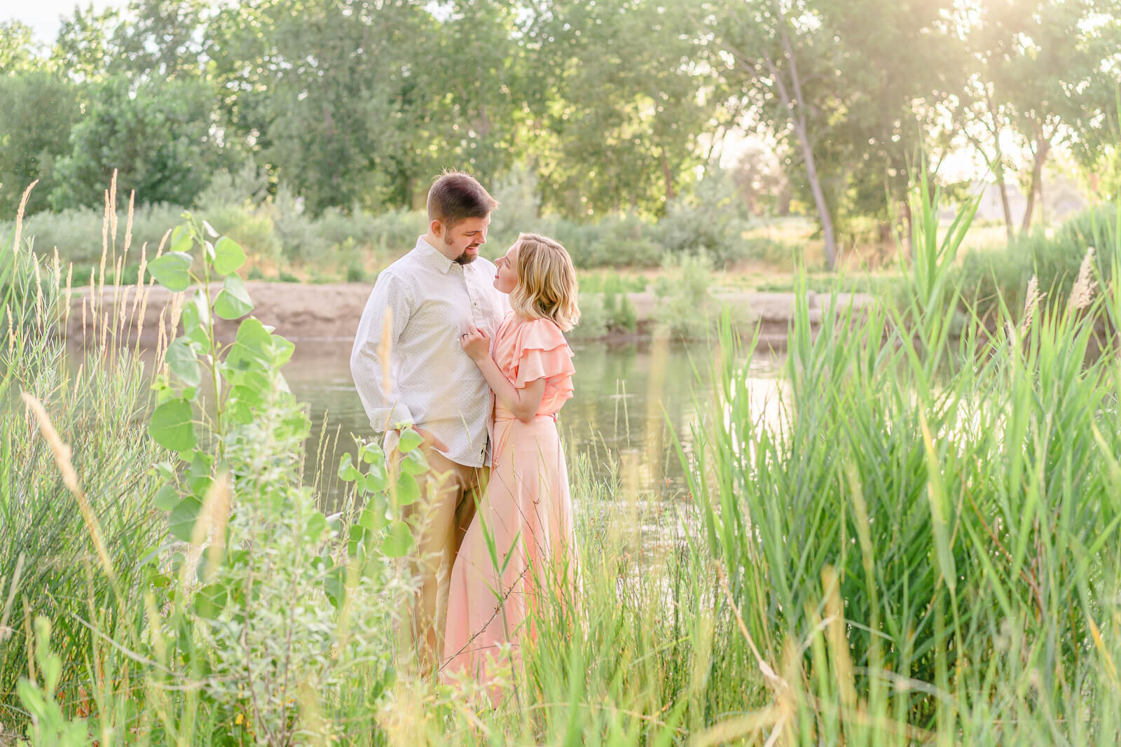 An engaged couple look at each other as they stand in front of a river at sunset
