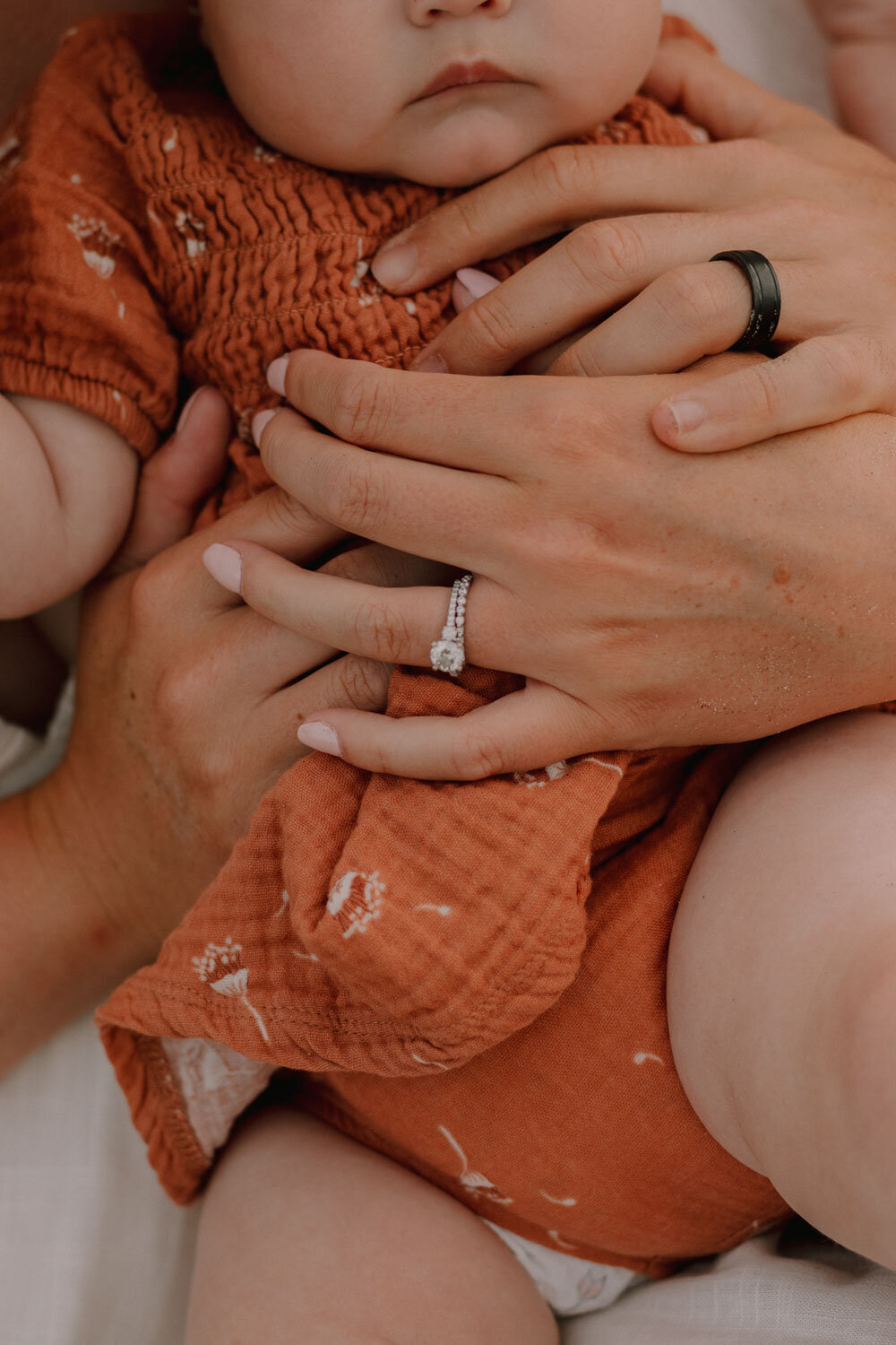Baby in orange outfit, hands held by parents with rings visible