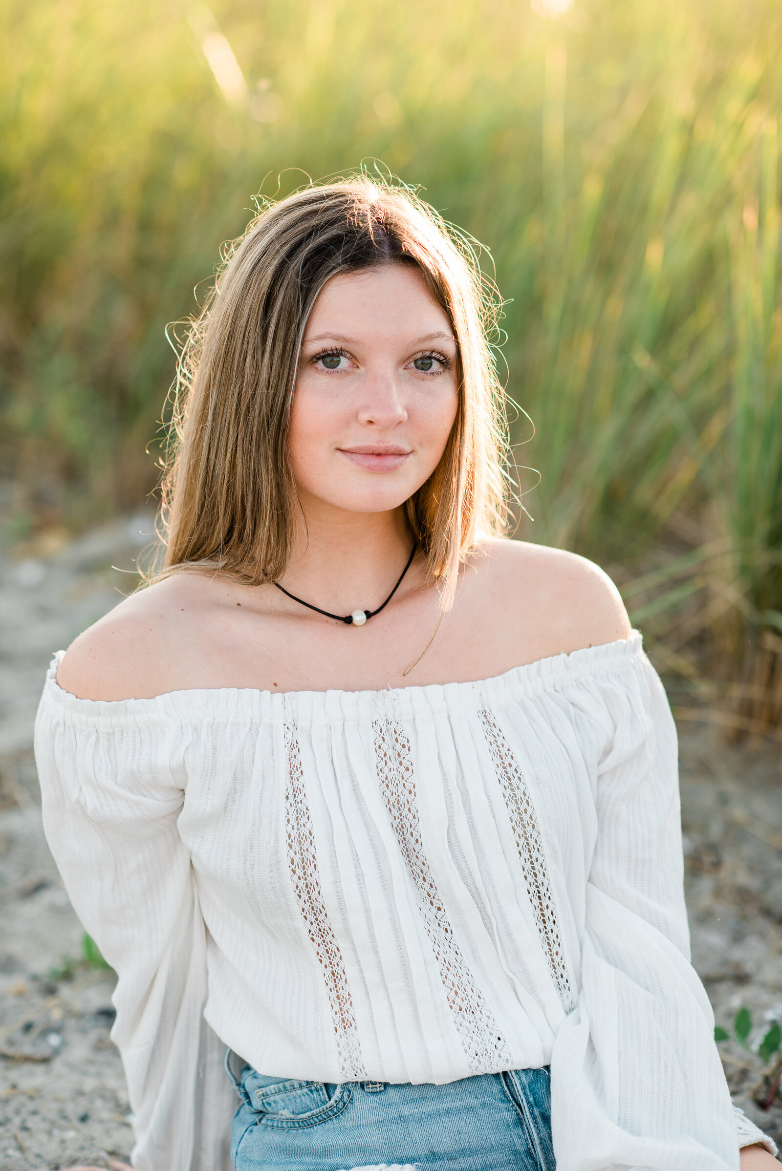 senior girl in sunlight sitting on sand in front of long grass