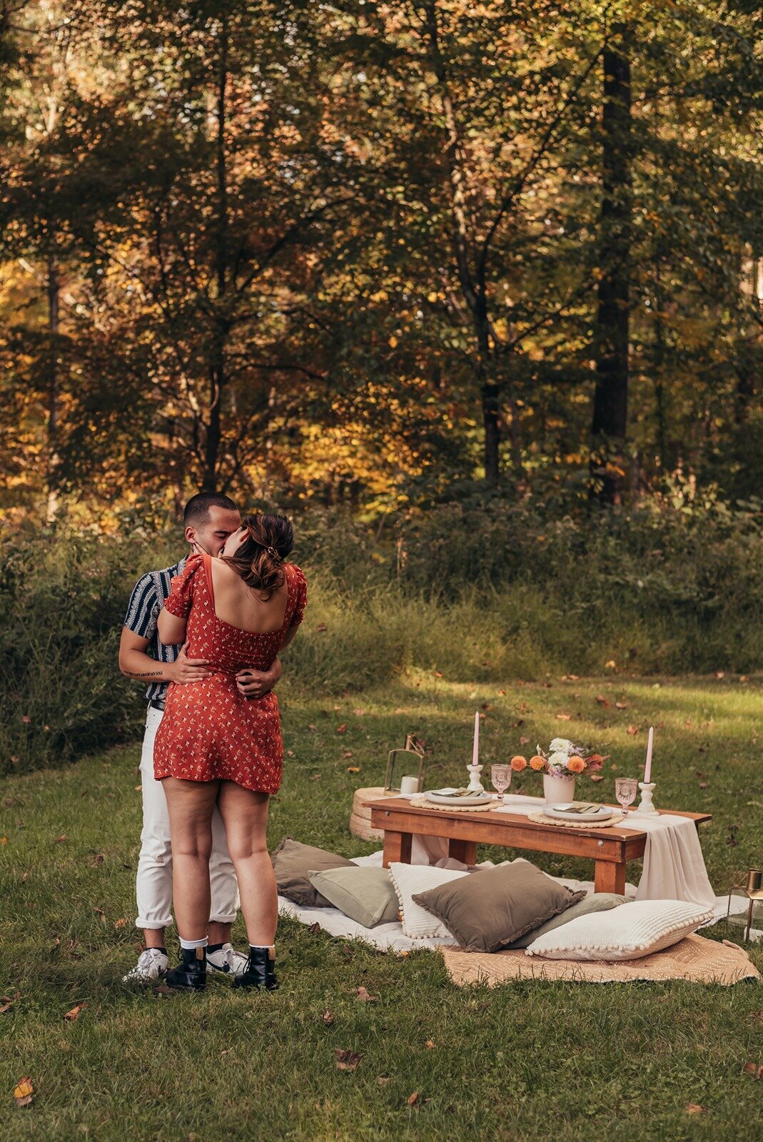 Couple embracing in front of a romantic picnic table with candles, florals, and cushions in a wooded outdoor setting, styled by Yours Truly, a wedding planner and designer in New Jersey