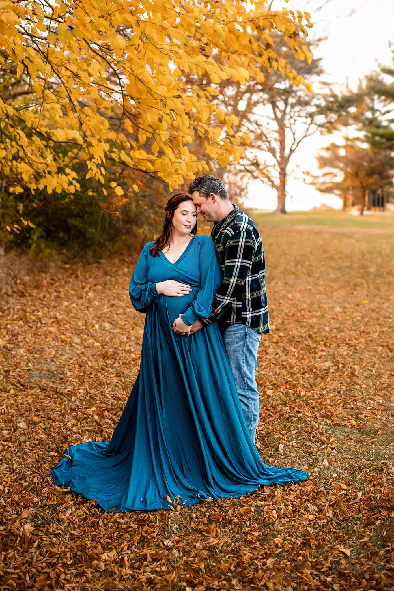 A pregnant woman in a flowing teal dress stands on autumn leaves, cradling her belly, while a man in a plaid shirt embraces her from behind. Golden trees frame this heartfelt moment, perfect for maternity portraits by a Bloomington maternity photographer.